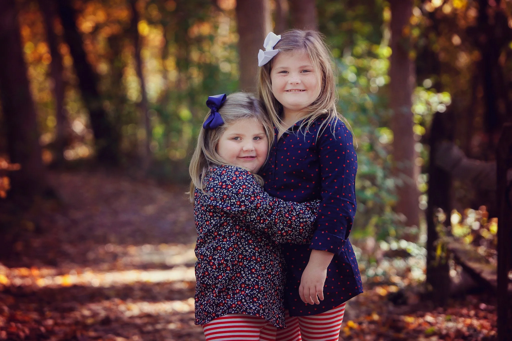 Two young girls standing close together outdoors in a wooded area during fall, both wearing dark blue jackets with red and white patterns and red and white striped leggings, smiling at the camera.