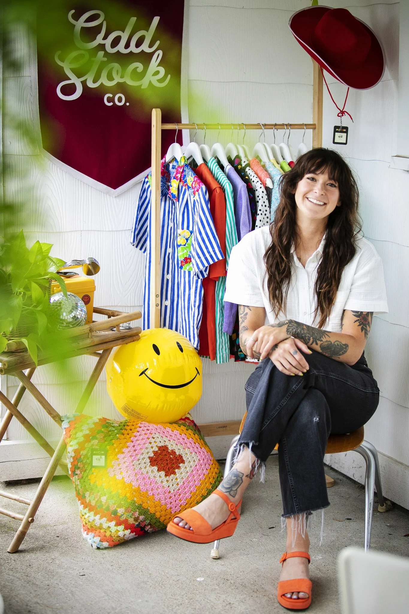 A woman sitting on a chair inside a clothing store with a rack of colorful shirts behind her. She is smiling, wearing a white shirt, ripped black jeans, and orange sandals. Beside her are a yellow smiley face balloon and a multicolored crochet pillow