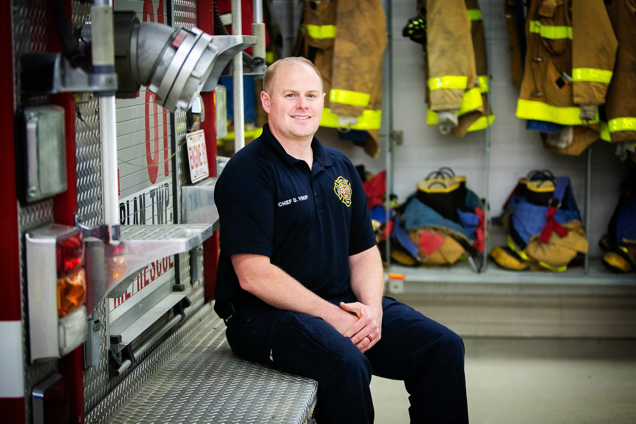 A firefighter in a navy uniform sitting on the edge of a fire truck inside a fire station, with firefighting gear hanging on the wall behind him.