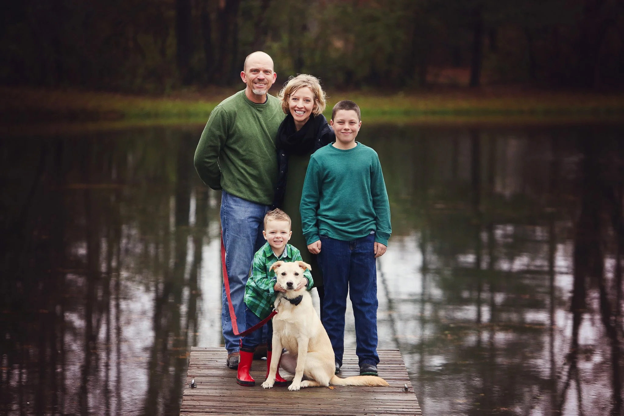 A family of four with a dog standing on a wooden dock by a lake in a wooded area during fall.