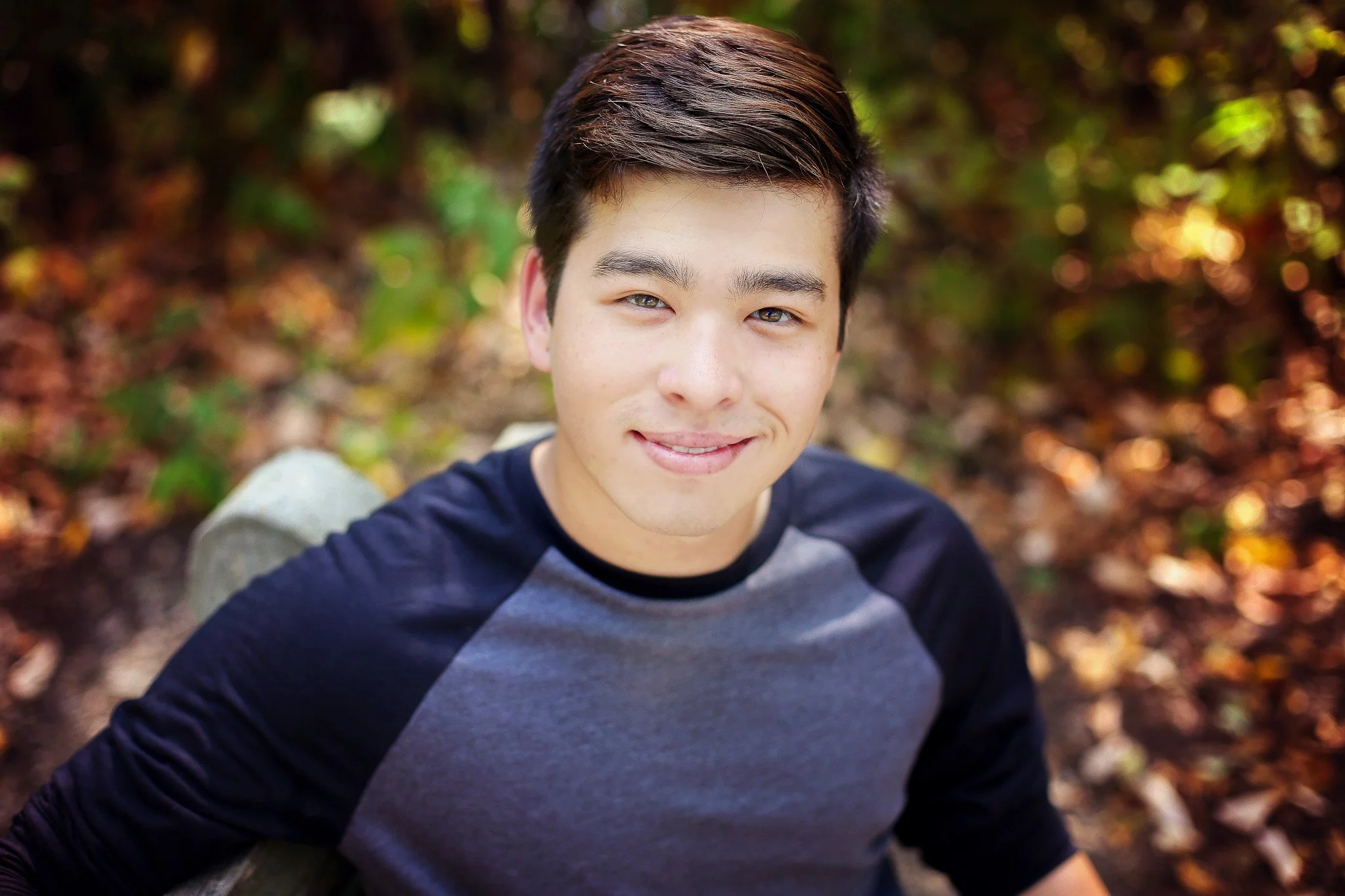 Young man with short dark hair and light skin, wearing a black and gray baseball tee, smiling outdoors with a blurred background of fall foliage.