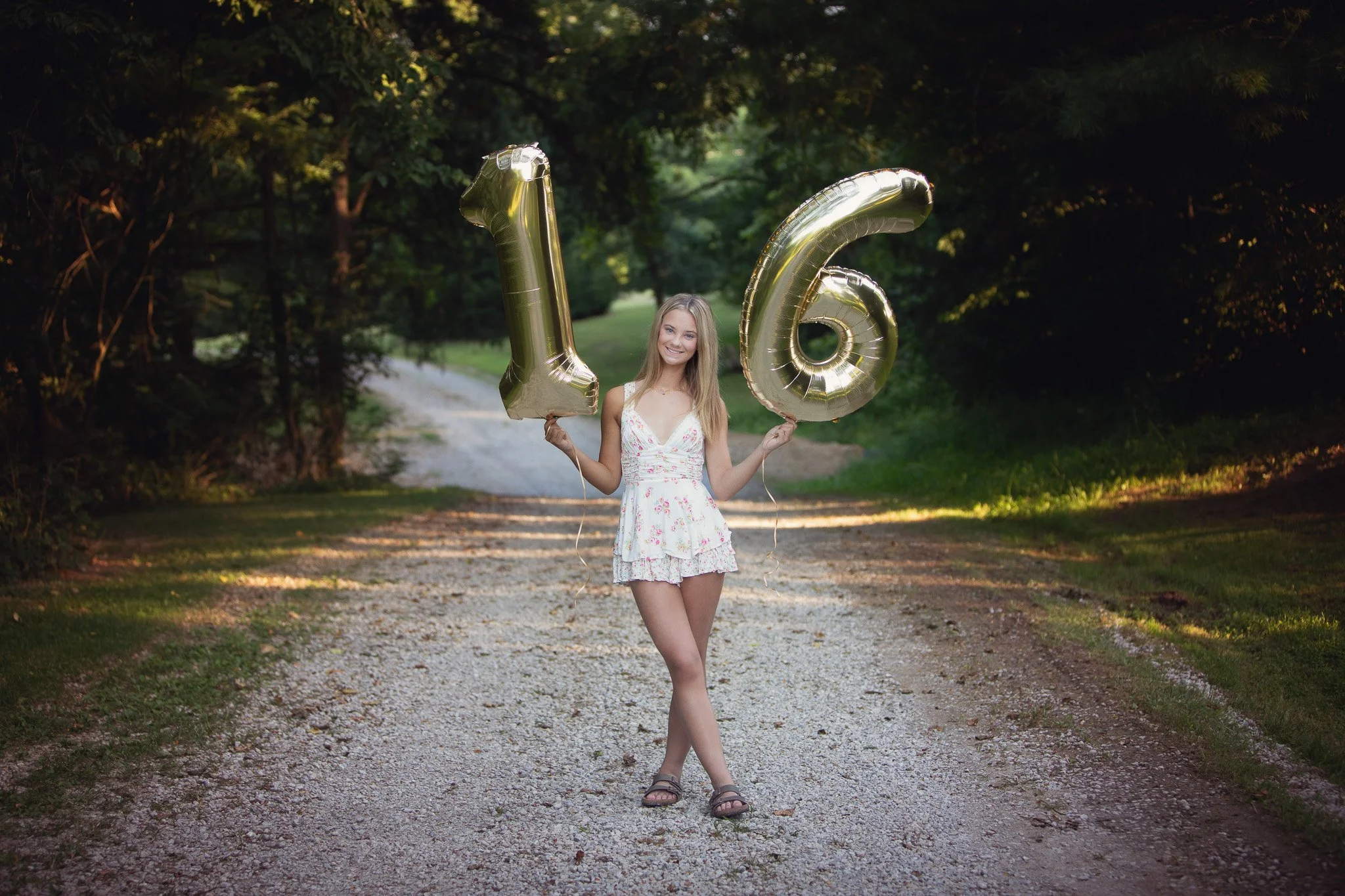 Young woman standing on a gravel path holding large, gold balloon numbers 1 and 6, celebrating her 16th birthday, smiling and wearing a white dress with pink floral pattern, surrounded by a wooded area.