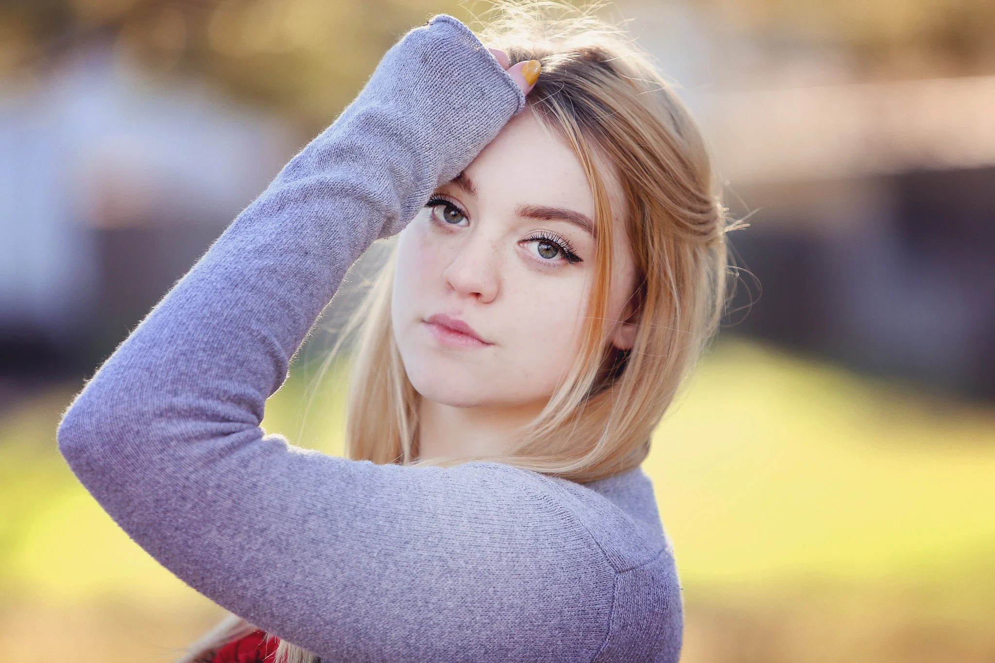 Portrait of a young woman with blonde hair and blue eyes, wearing a gray sweater, standing outdoors in a park with blurred autumn background.