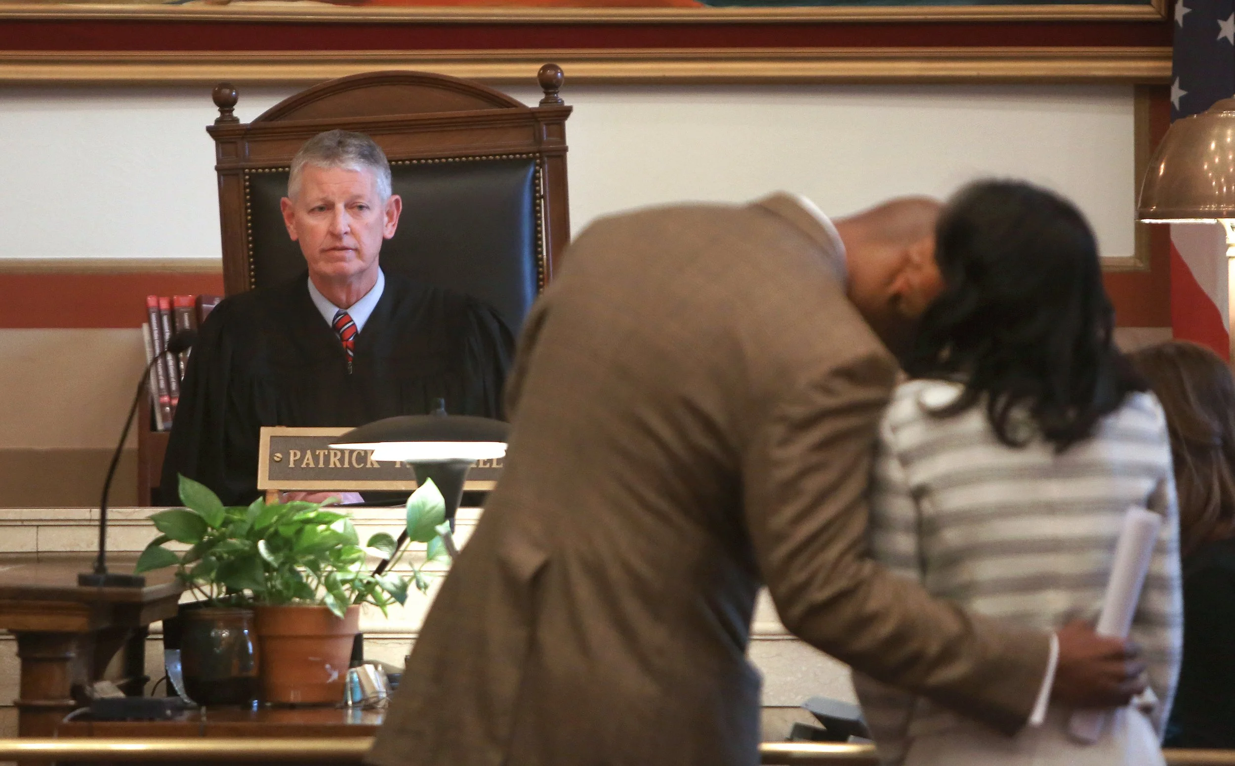 A judge in a courtroom courtroom, dressed in a black robe, sitting behind a desk with a nameplate that reads 'Patrick'. Two individuals are in the foreground, one leaning over to speak to another woman who is holding papers, and a potted plant is on 