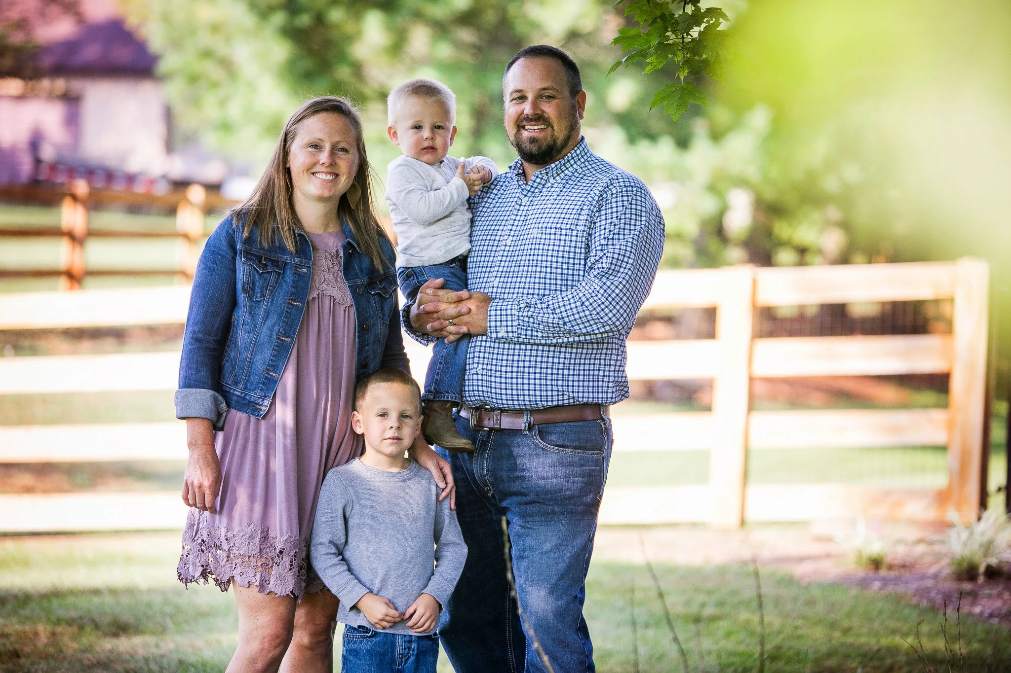 A family of five standing outdoors near a wooden fence, smiling. The mother, father, and three children (two boys and a toddler) are posing together on a sunny day with trees in the background.