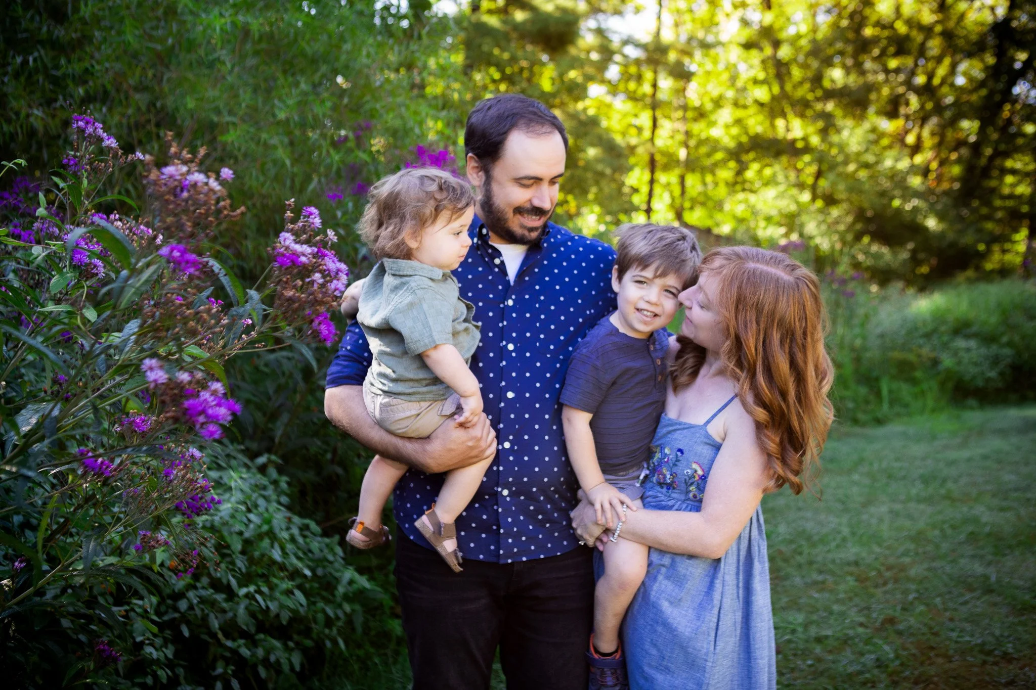A family of four standing on a grassy area next to purple flowers and green trees, smiling and enjoying time together outdoors.