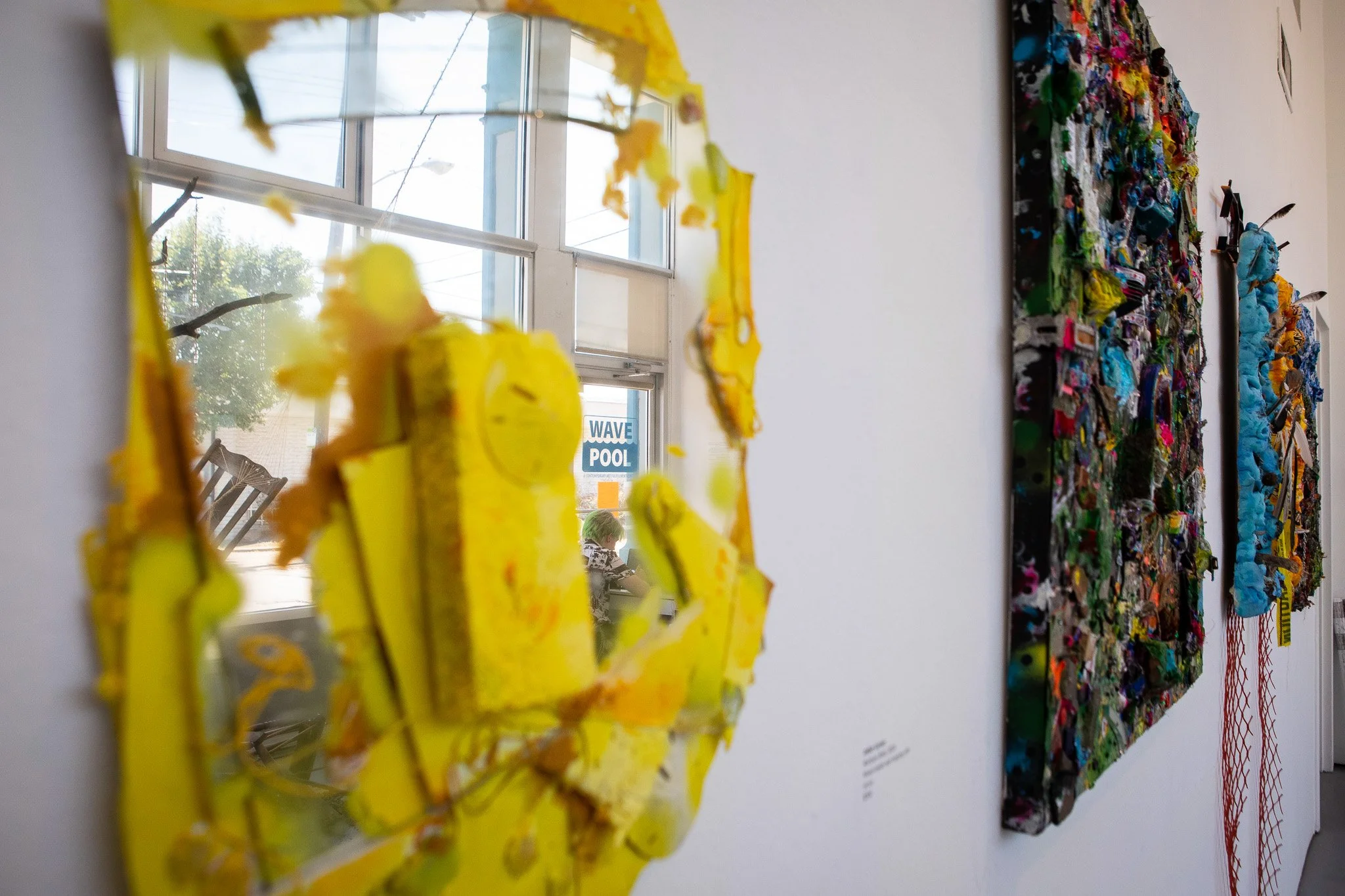 Close-up of a mirror with irregular yellow edges on a white wall, reflecting a window and a person sitting at a table.