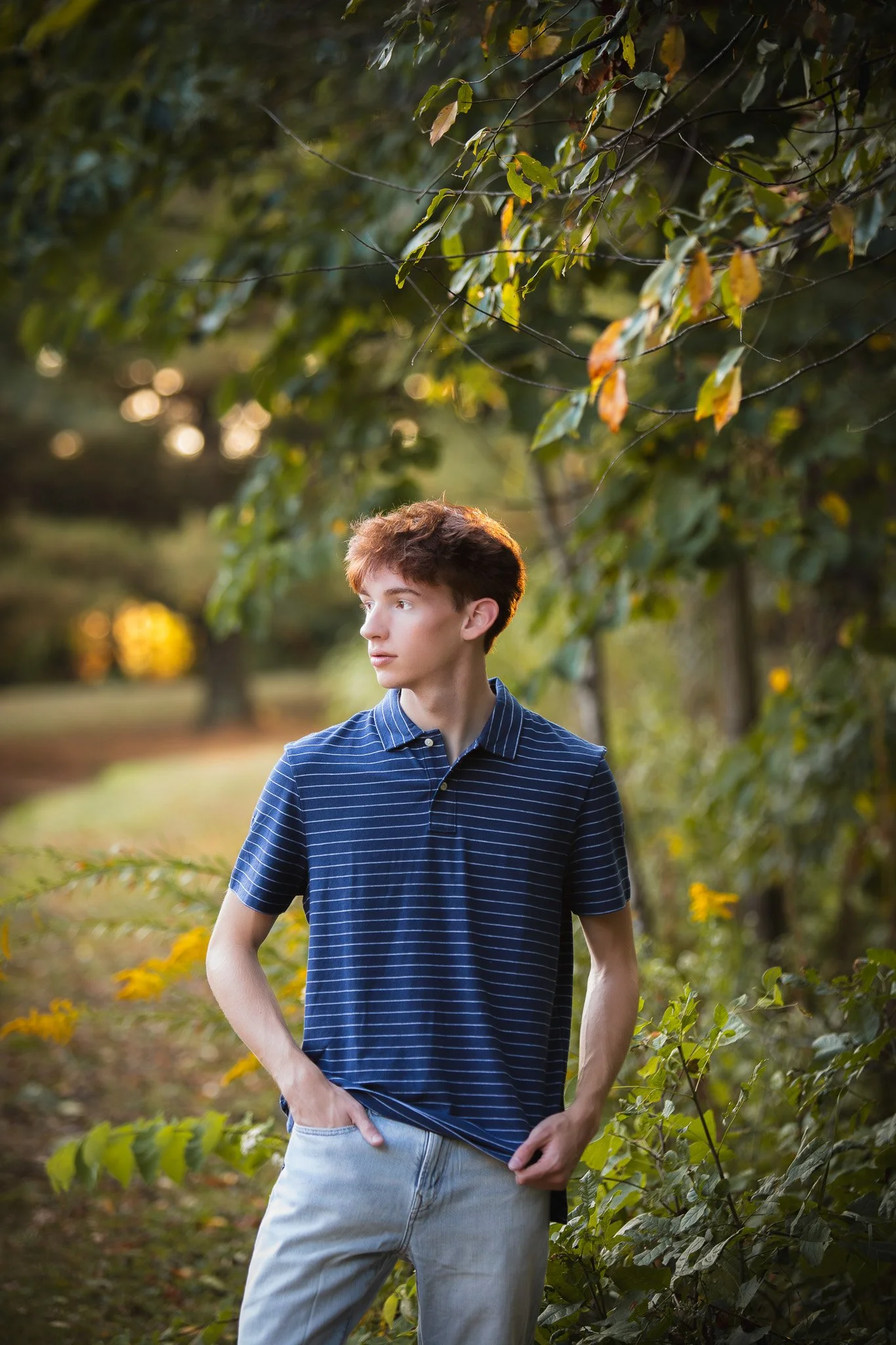A young man with curly brown hair wearing a blue and white striped polo shirt and light jeans standing outdoors in a park with greenery and trees.