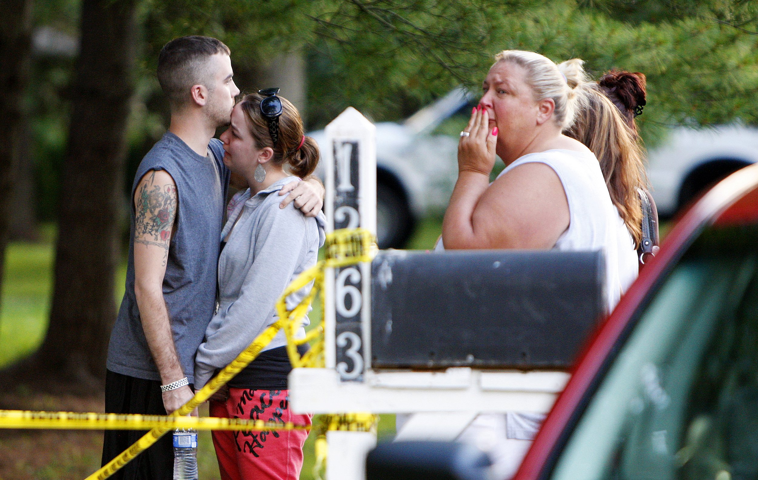 A young man and woman embrace and share a kiss while a woman nearby reacts with shock, covering her mouth with her hand. The scene appears to be outdoors in a park or similar setting.