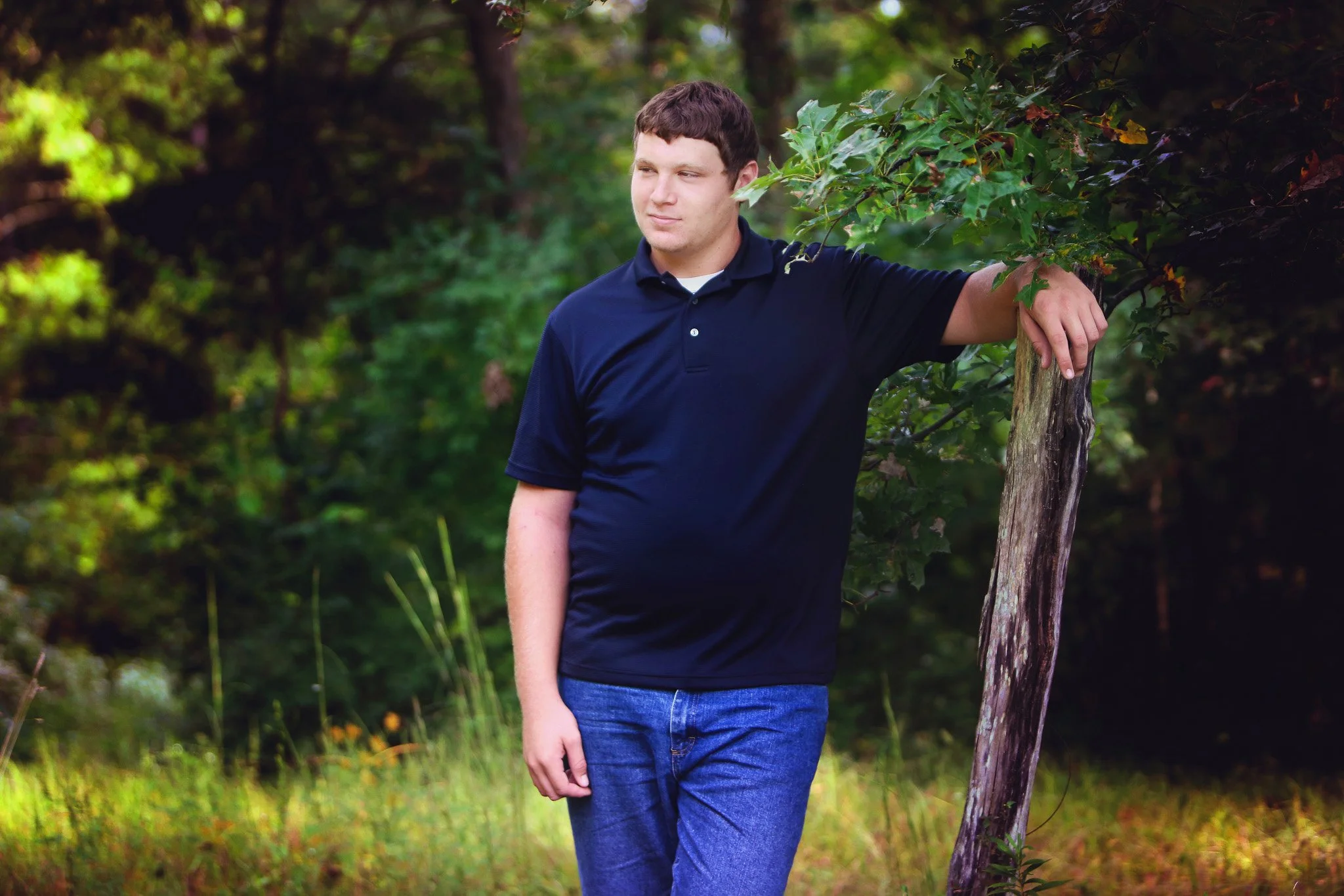 Young man leaning on a tree in a forested area with lush green foliage.
