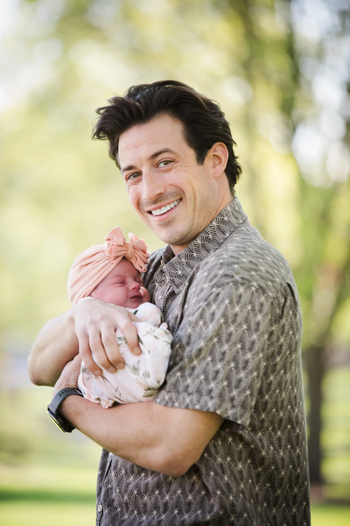 A man holding a newborn baby outdoors in a park with trees in the background. The man is smiling and the baby is wearing a pink headband.