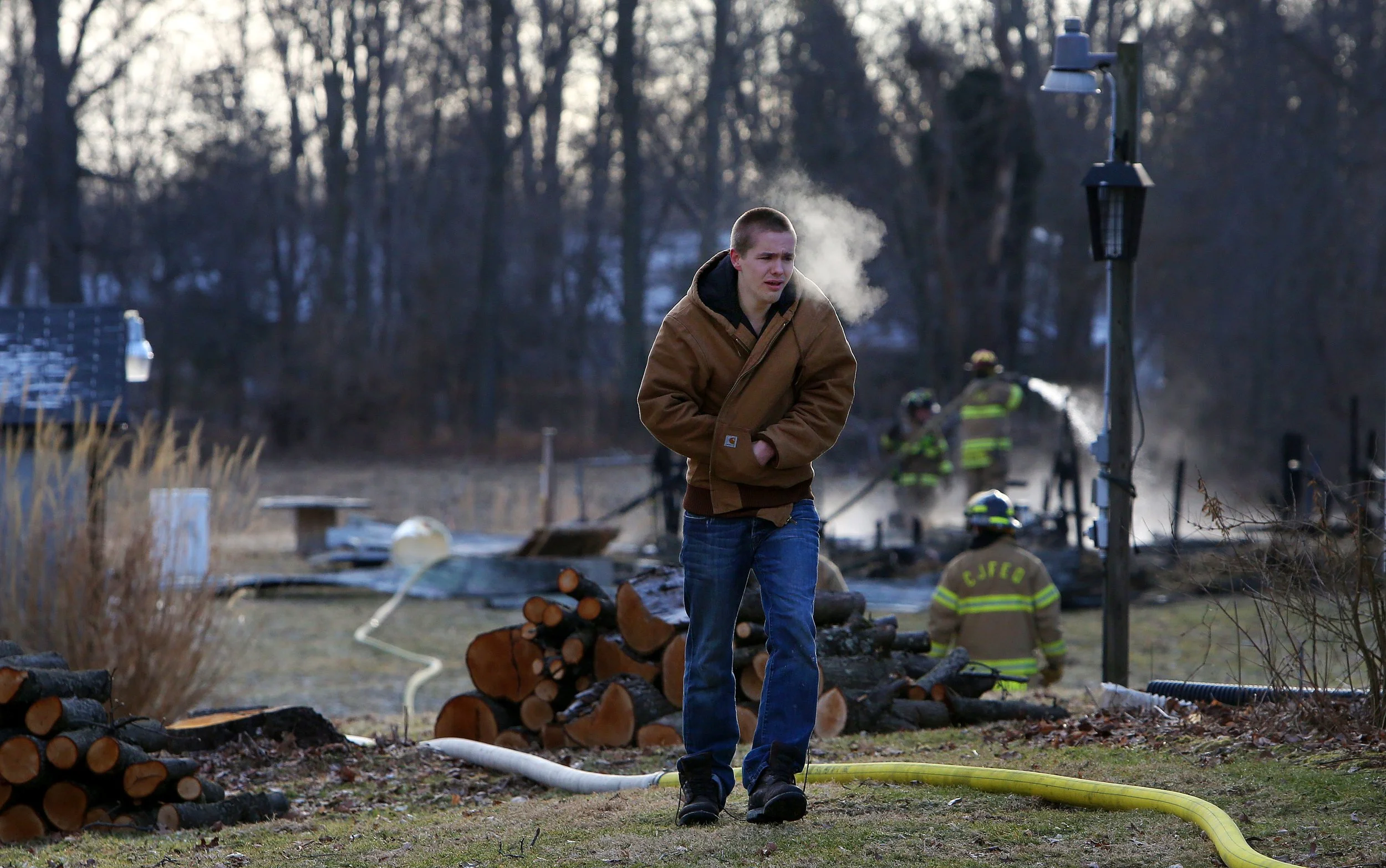 A young man walking away from a fire scene with firefighters working in the background, smoke rising from the area, logs on the ground, and a lamp post.
