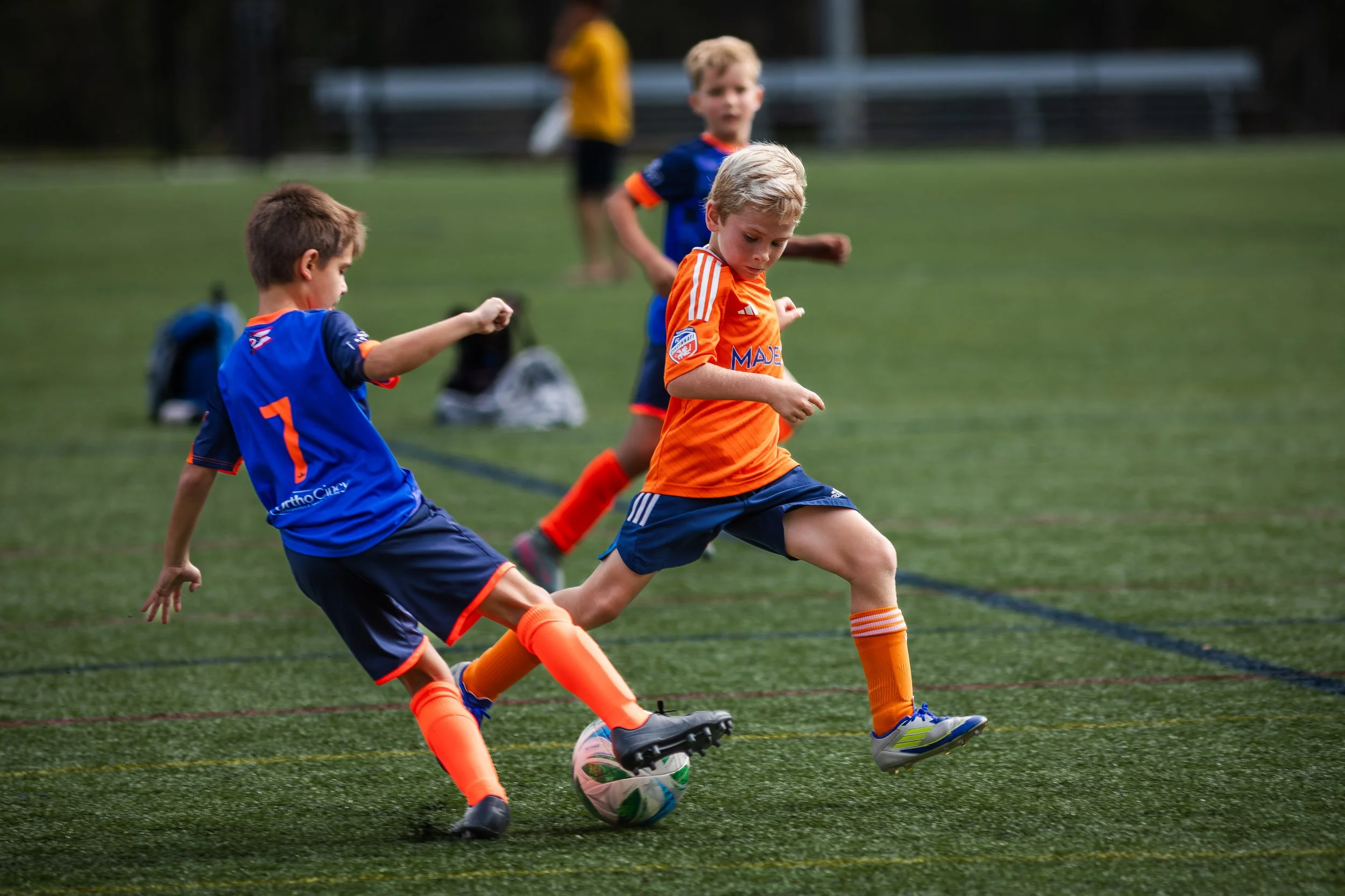 Kids playing soccer on a field, actively kicking the ball while two children in blue and orange uniforms compete
