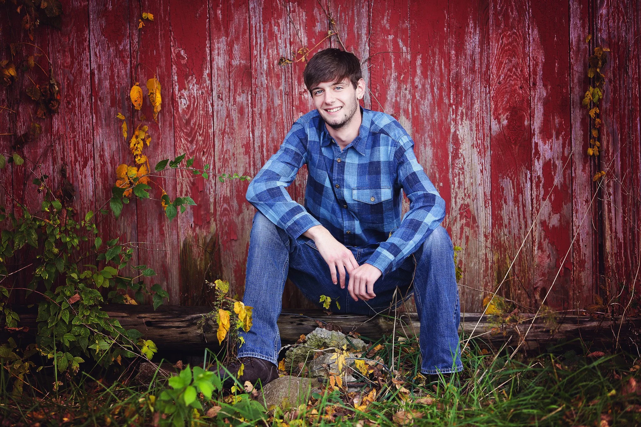 A young man sitting outdoors on a log in front of a weathered red wooden fence, wearing a blue plaid shirt and jeans, smiling and looking at the camera.