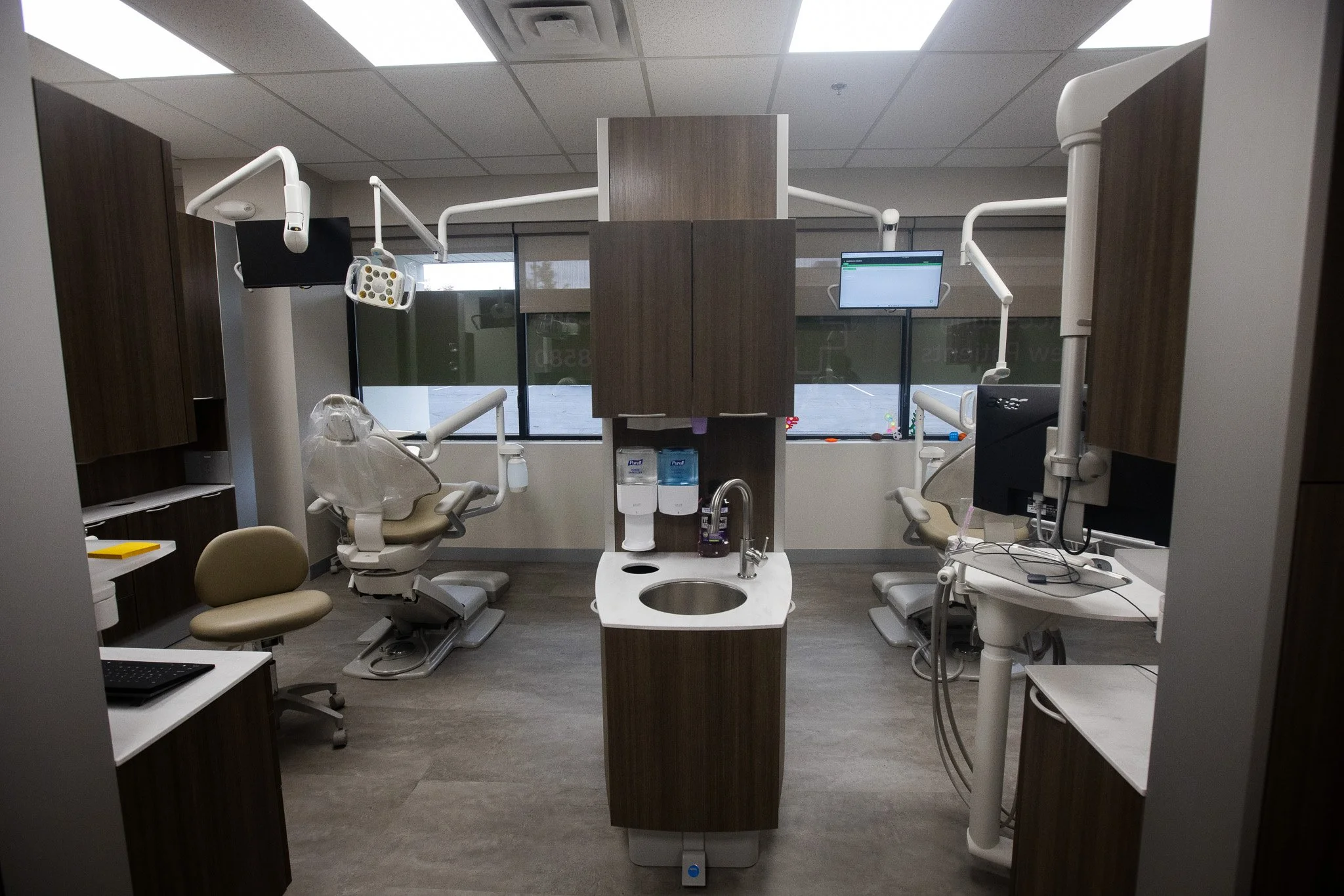 Modern dental office with two dental chairs, equipment, and monitors, seen through a doorway.
