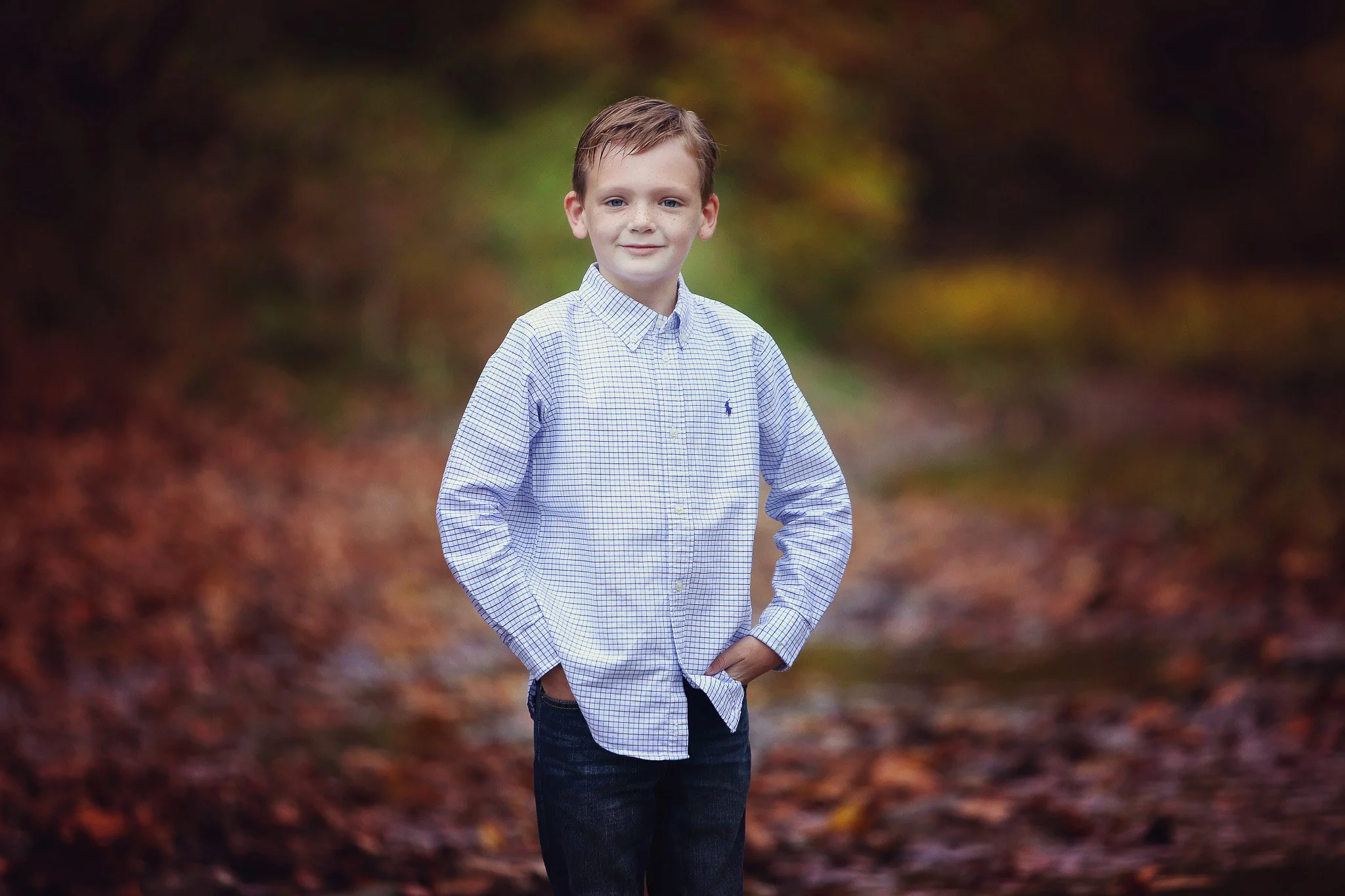 A young boy with brown hair and blue eyes, wearing a light blue checkered shirt and dark jeans, standing outdoors on a fall day with autumn leaves on the ground and blurred trees in the background.