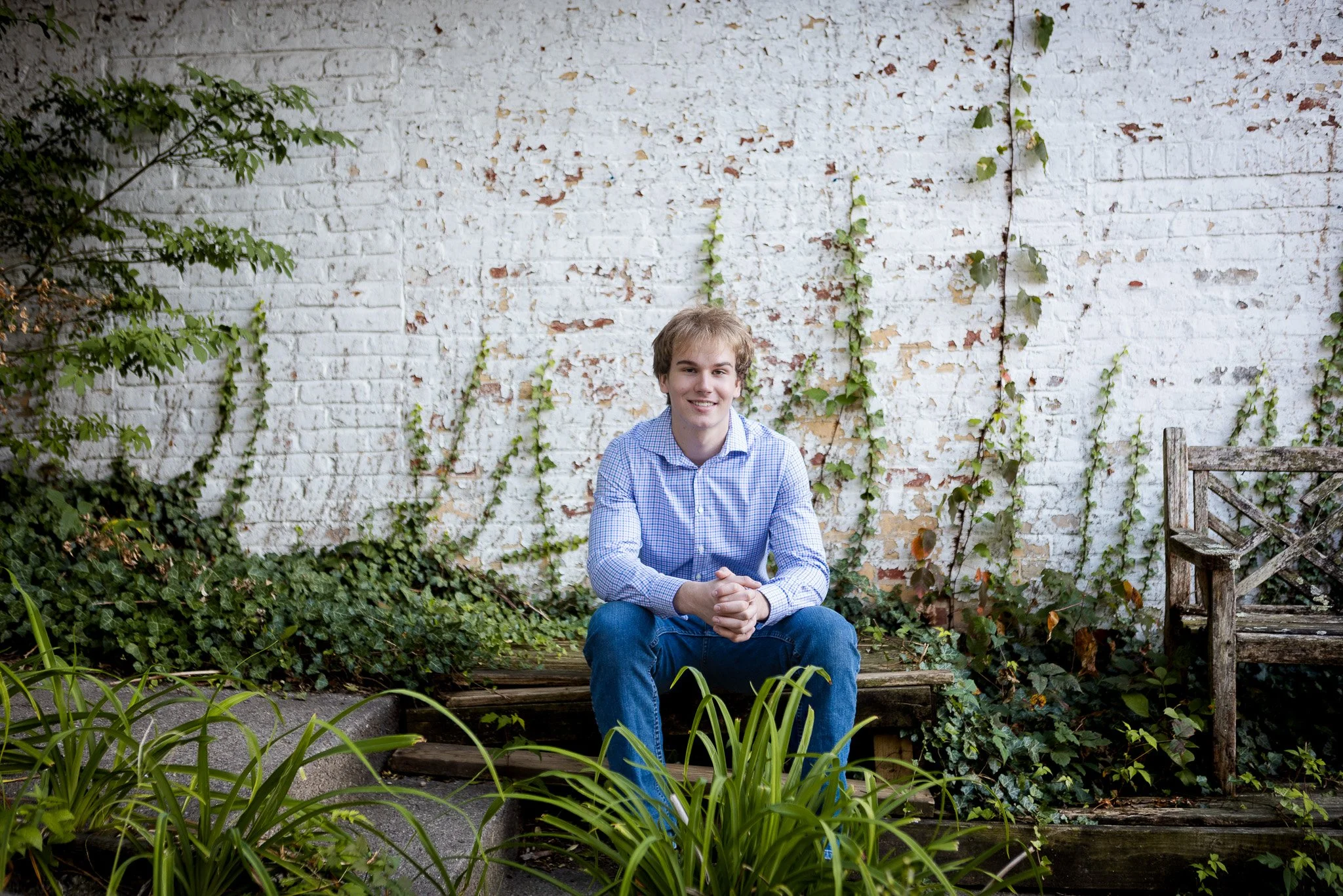 A young man in a blue checkered shirt and jeans sitting on a wooden bench outdoors, against a white brick wall with climbing ivy, surrounded by green plants.