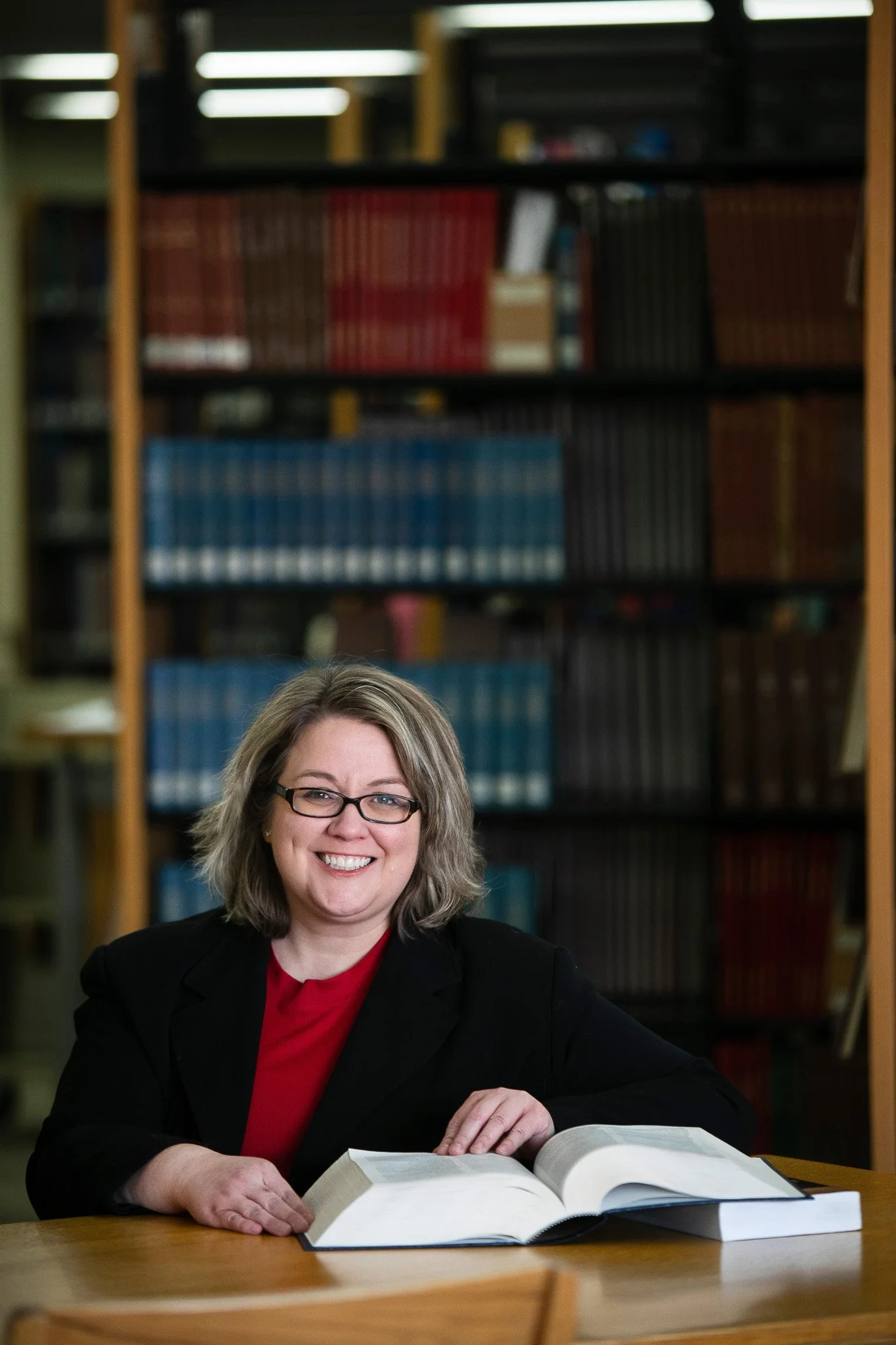 Woman with glasses smiling, sitting at a desk with an open book, in a library with bookshelves in the background.