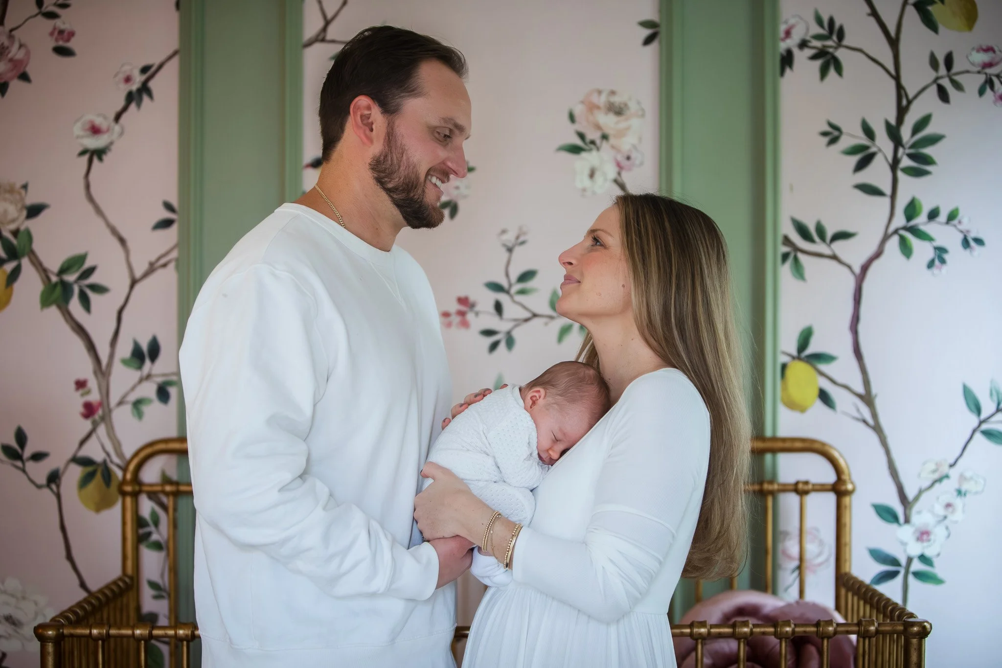 A couple holding a baby girl in a nursery with floral wall decor, the woman in a white dress and the man in a white shirt, both smiling at each other.
