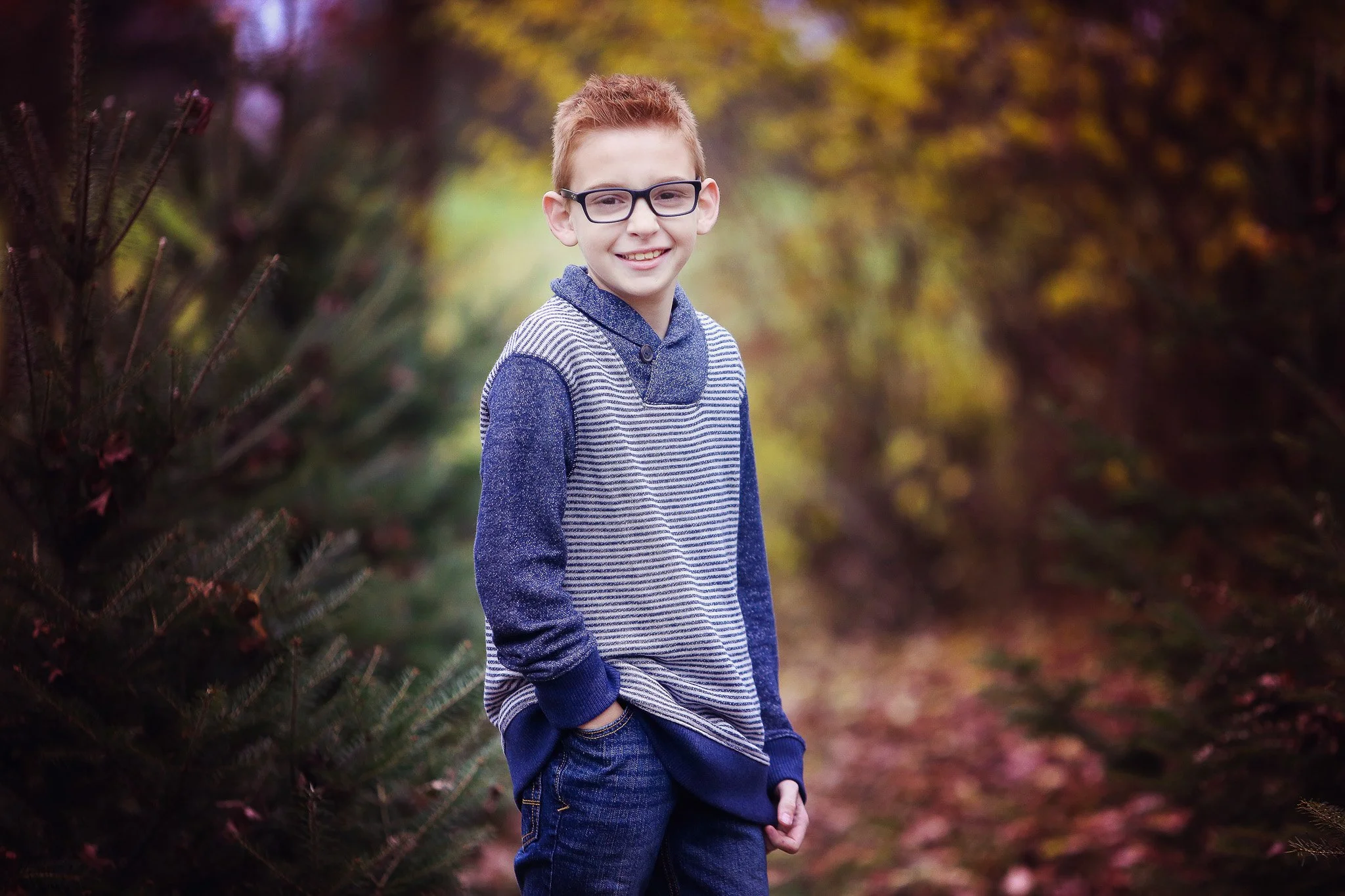A young boy with glasses and red hair smiling outdoors in a path surrounded by trees with autumn leaves.