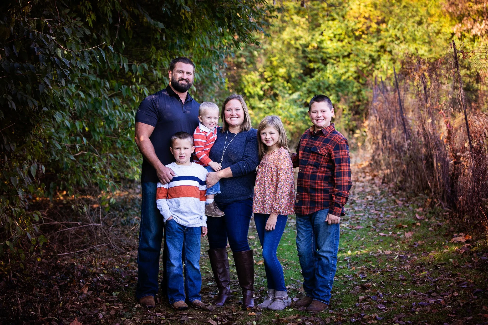 A family of six standing outdoors on a leaf-covered path with green and multicolored foliage in the background, smiling at the camera.