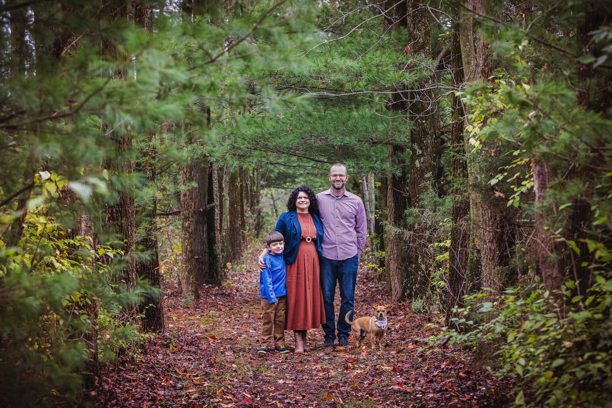 Family of three with dog standing on forest trail surrounded by evergreen trees.