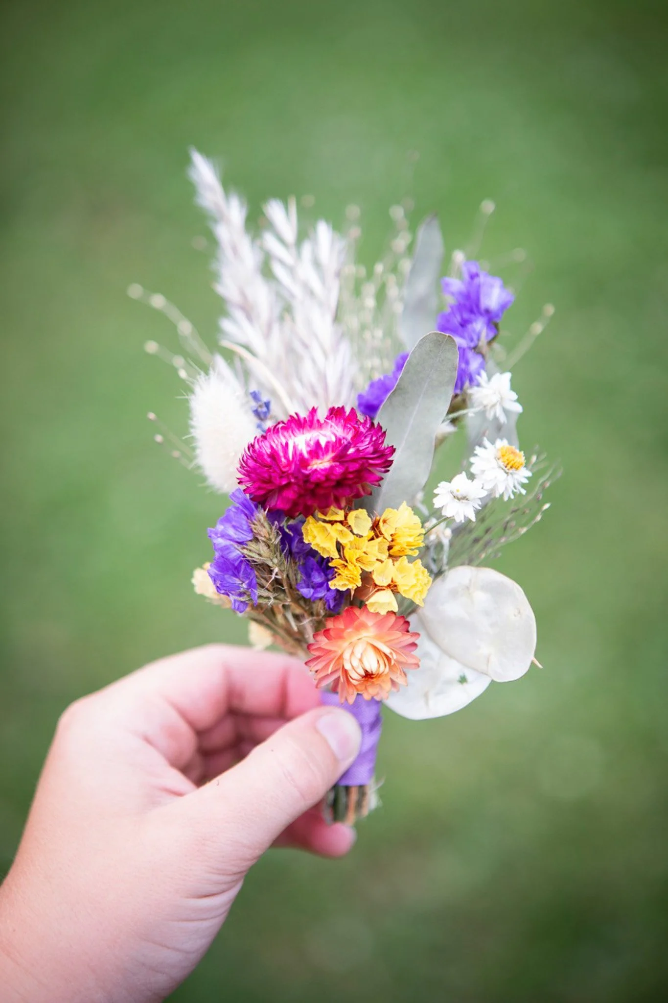 A hand holding a small bouquet of colorful dried flowers and leaves against a blurred green background.