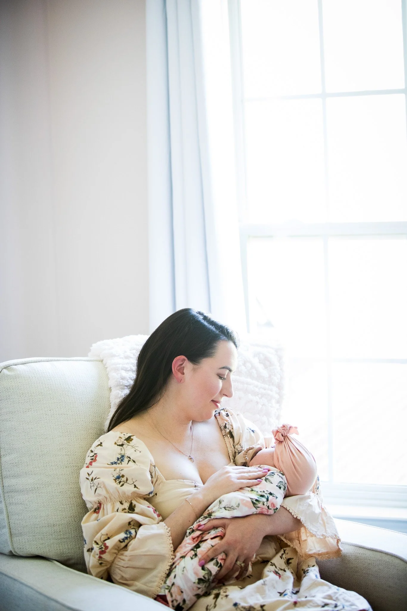 A woman with dark hair, wearing a floral dress, sitting on a beige armchair, breastfeeding a baby in a pink outfit, near a large window with white curtains.