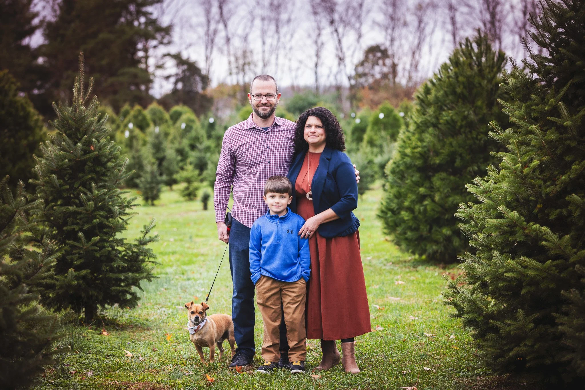A family of three with a dog standing in a Christmas tree farm with evergreen trees in the background.