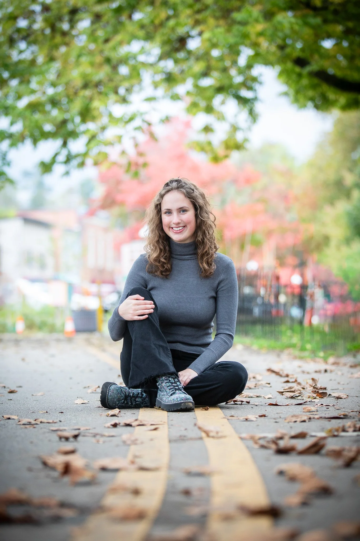 A young woman smiling and sitting cross-legged on a street with fallen leaves, surrounded by autumn trees.
