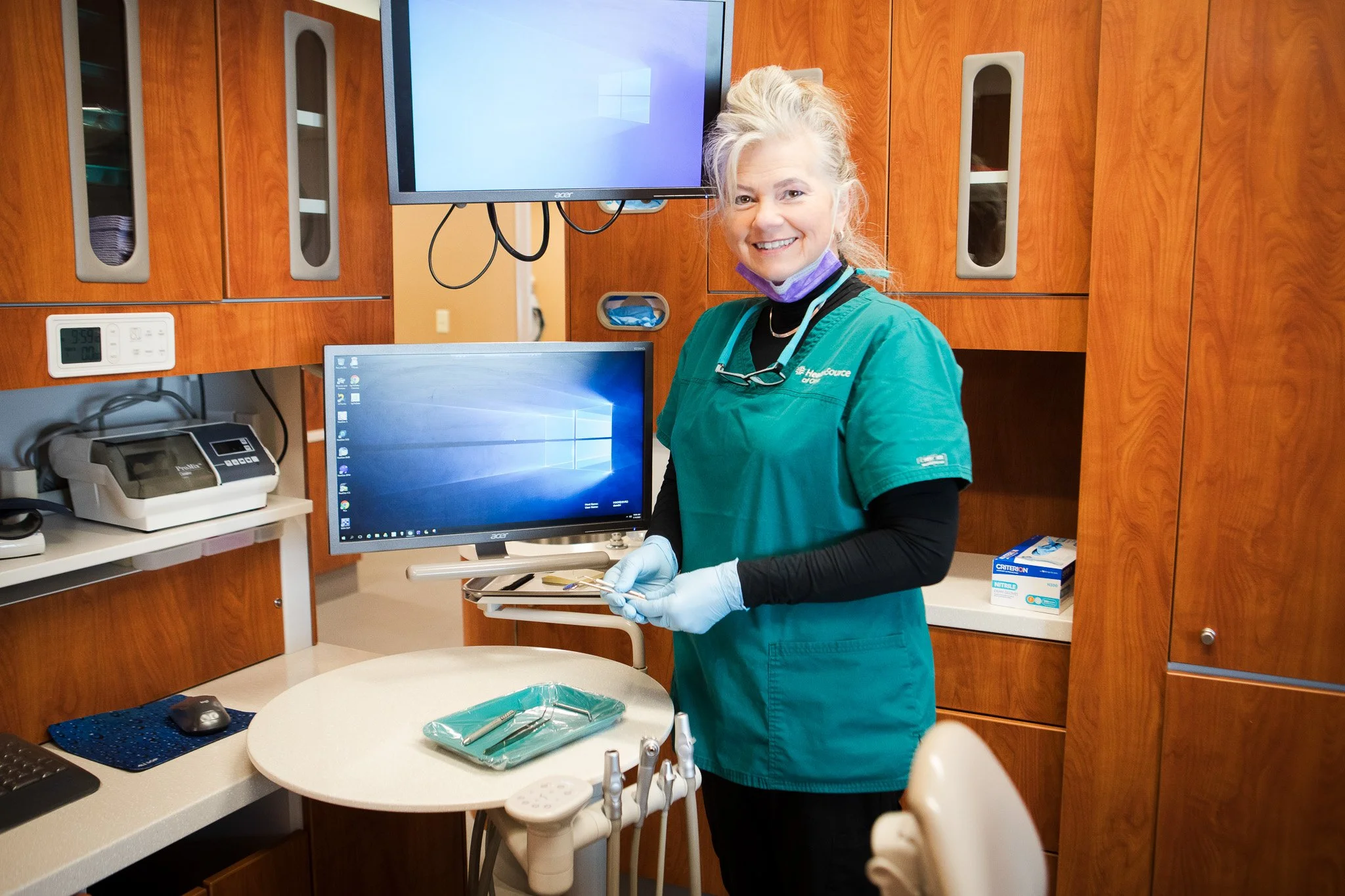 A dental professional wearing teal scrubs, gloves, and a mask around her neck, standing in a clinical setting, smiling at the camera, with computer monitors and dental equipment visible in the background.
