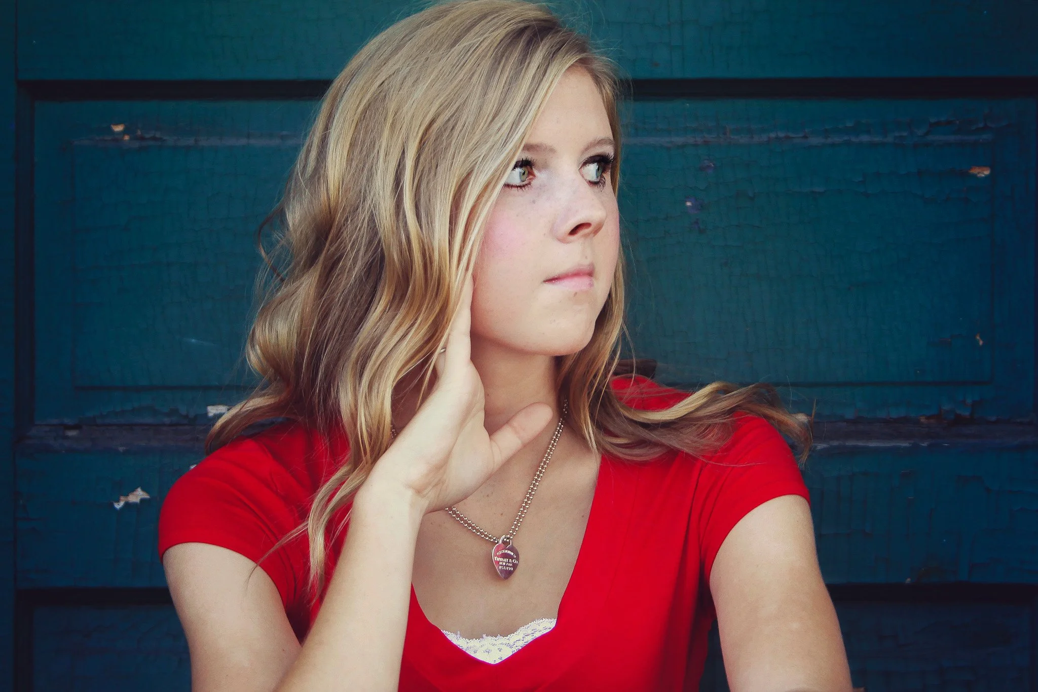 A young woman with long blonde hair wearing a red shirt and a heart-shaped necklace, looking to her left with her hand resting on the side of her face, standing in front of a blue textured background.