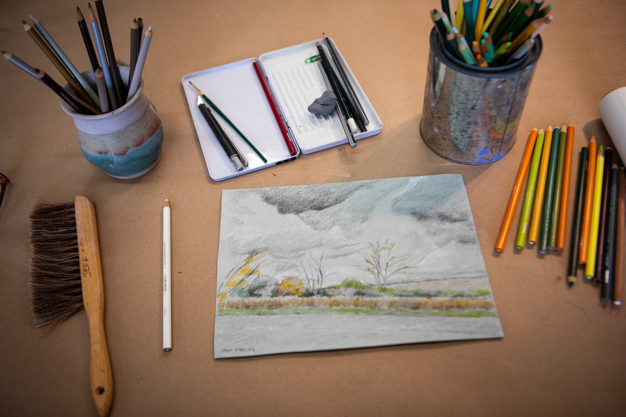 Art supplies including colored pencils, watercolor paints, brushes, and a drawing of a landscape on paper are arranged on a table.