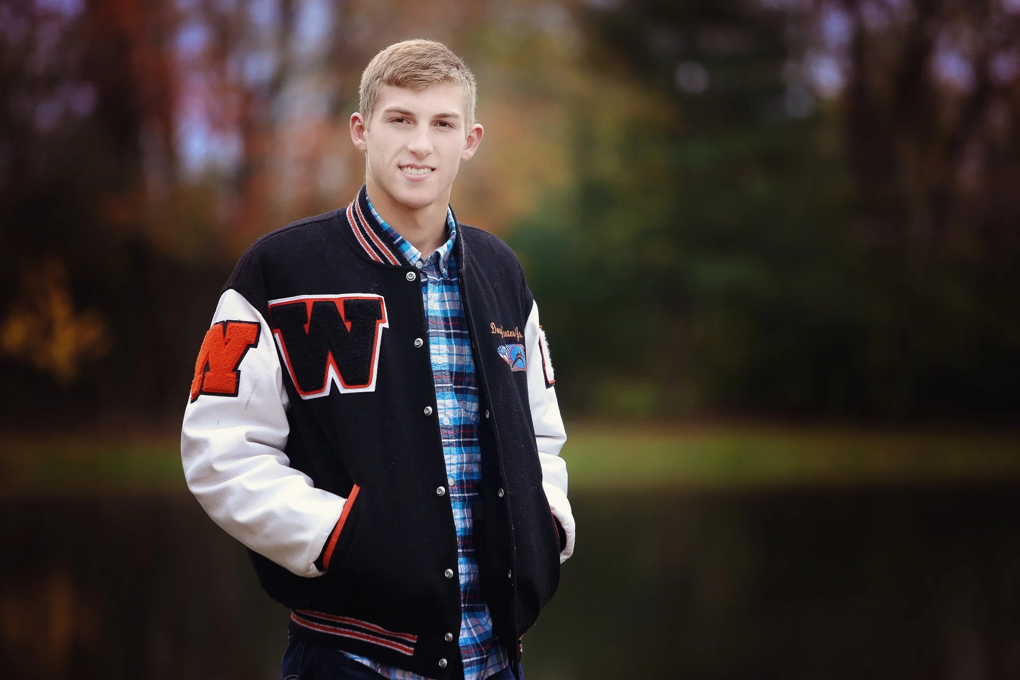 A young man standing outdoors near a body of water in autumn, wearing a black letterman jacket with white sleeves, a blue plaid shirt, and jeans.