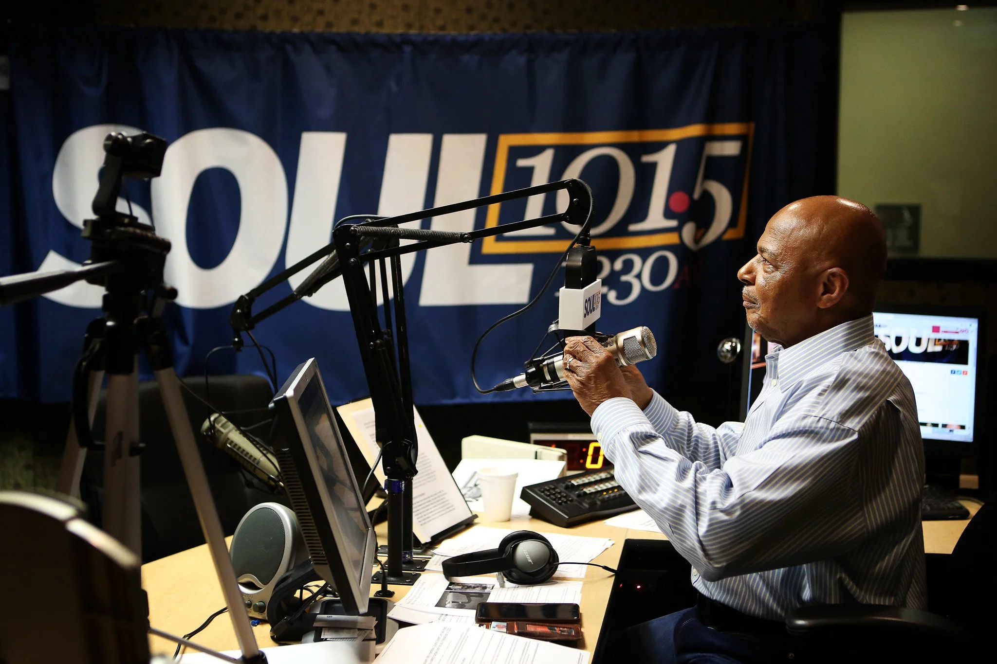 Man speaking into a microphone in a radio studio with a large political campaign banner in the background.