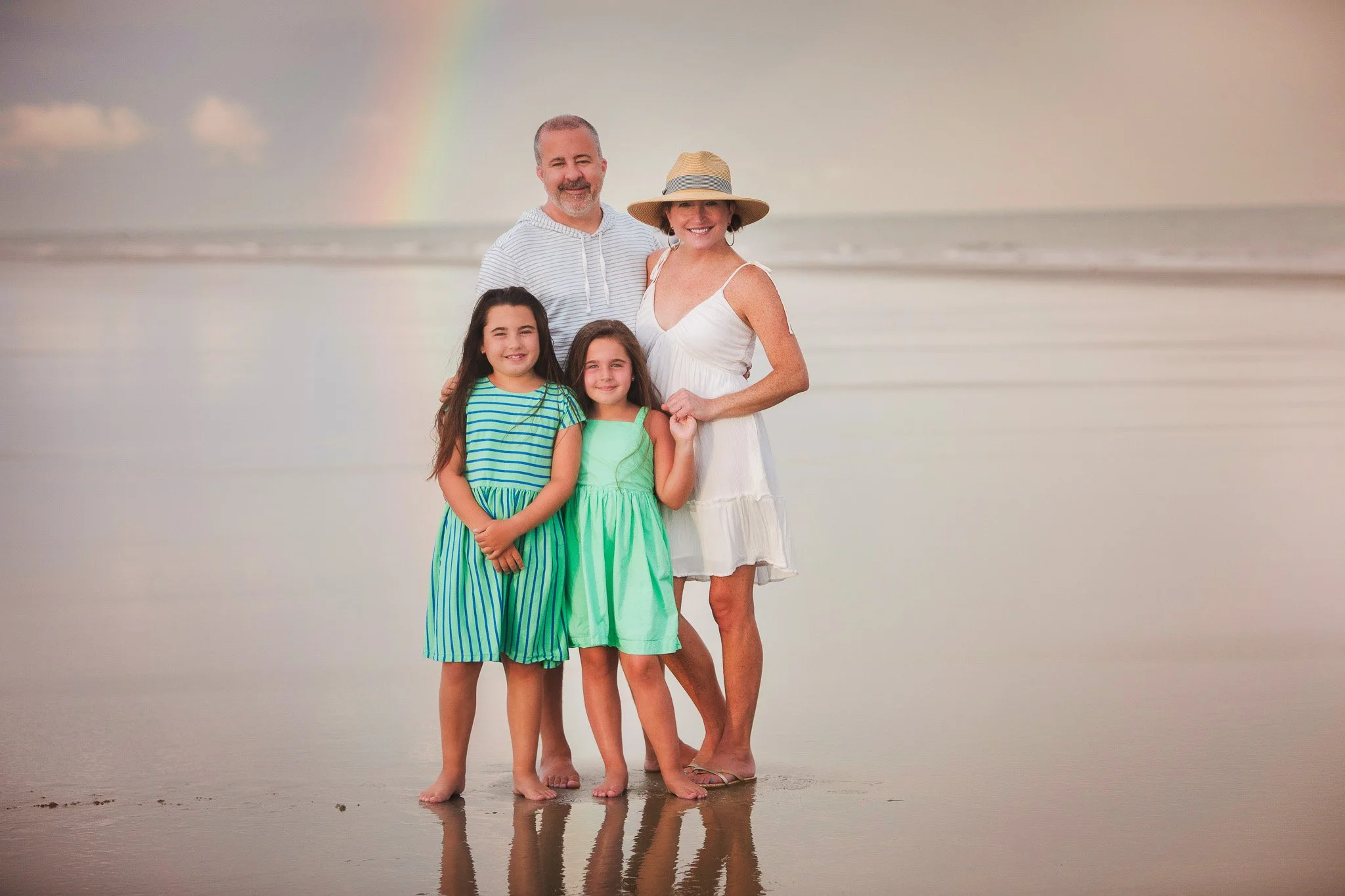 A family of four standing on the beach with a rainbow in the background, the ocean behind them.