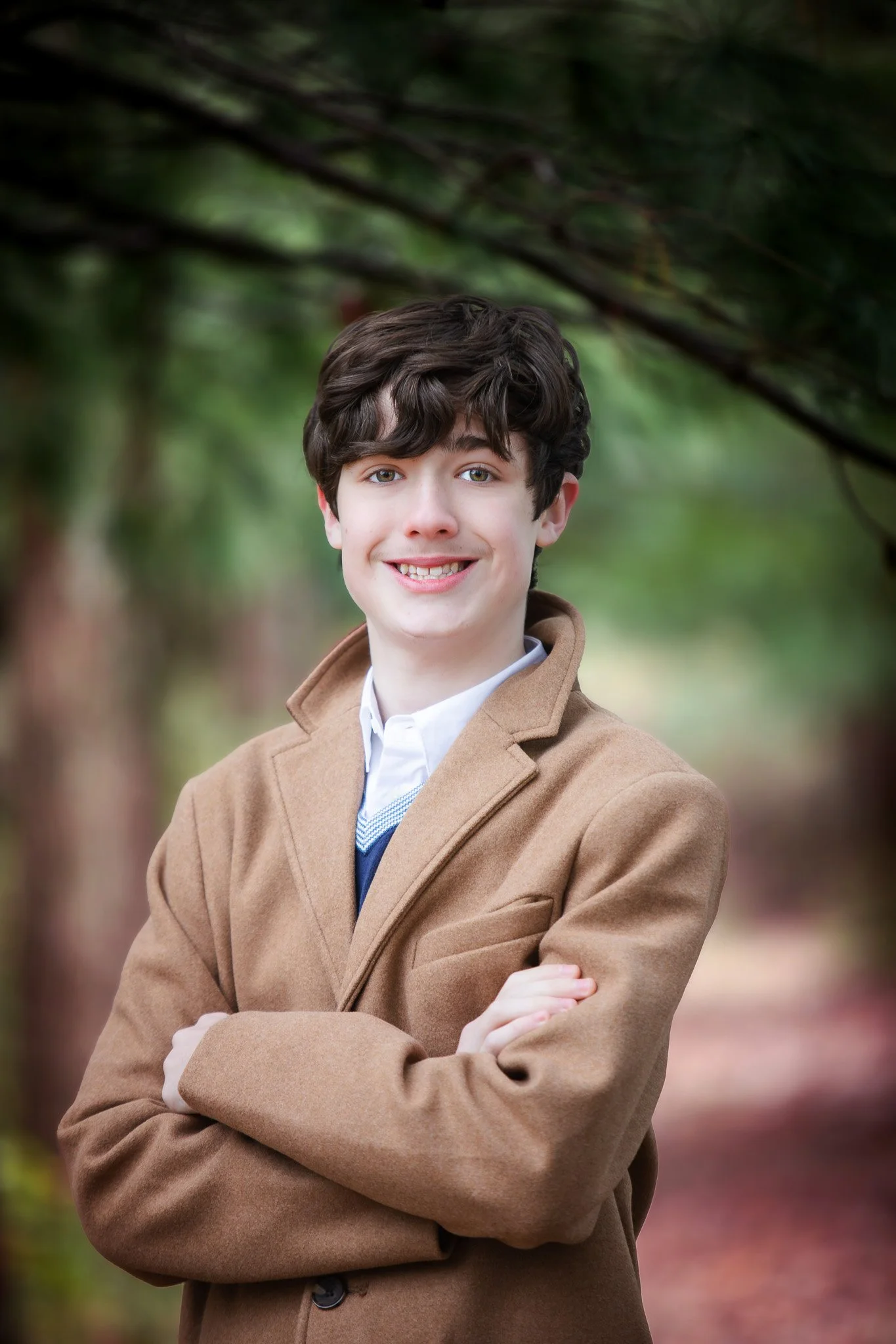 A young man standing outdoors in front of green trees, smiling with arms crossed, wearing a tan coat over a white shirt and a sweater.
