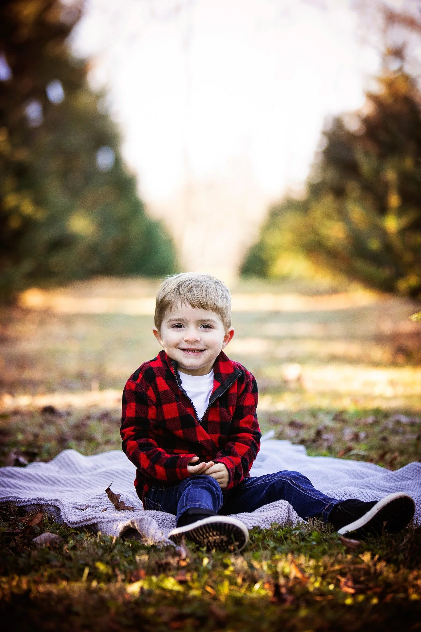 A young boy sitting on a blanket outdoors in a park during fall, smiling at the camera, wearing a red and black plaid jacket, white shirt, and jeans.