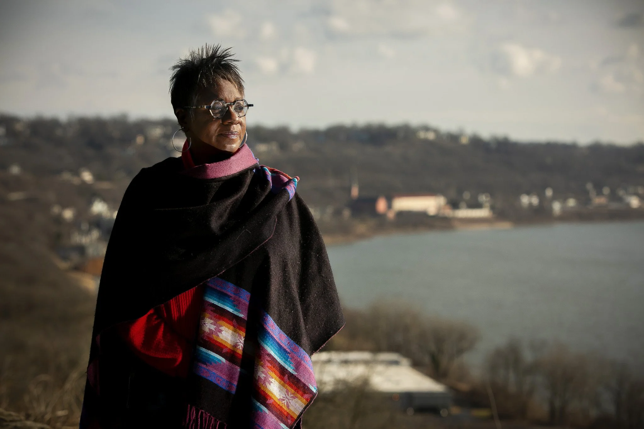 A woman standing outdoors near a body of water, looking to the side. She has short dark hair, glasses, and is wearing a black shawl with colorful patterns and a pink scarf. The background shows hills and some buildings.