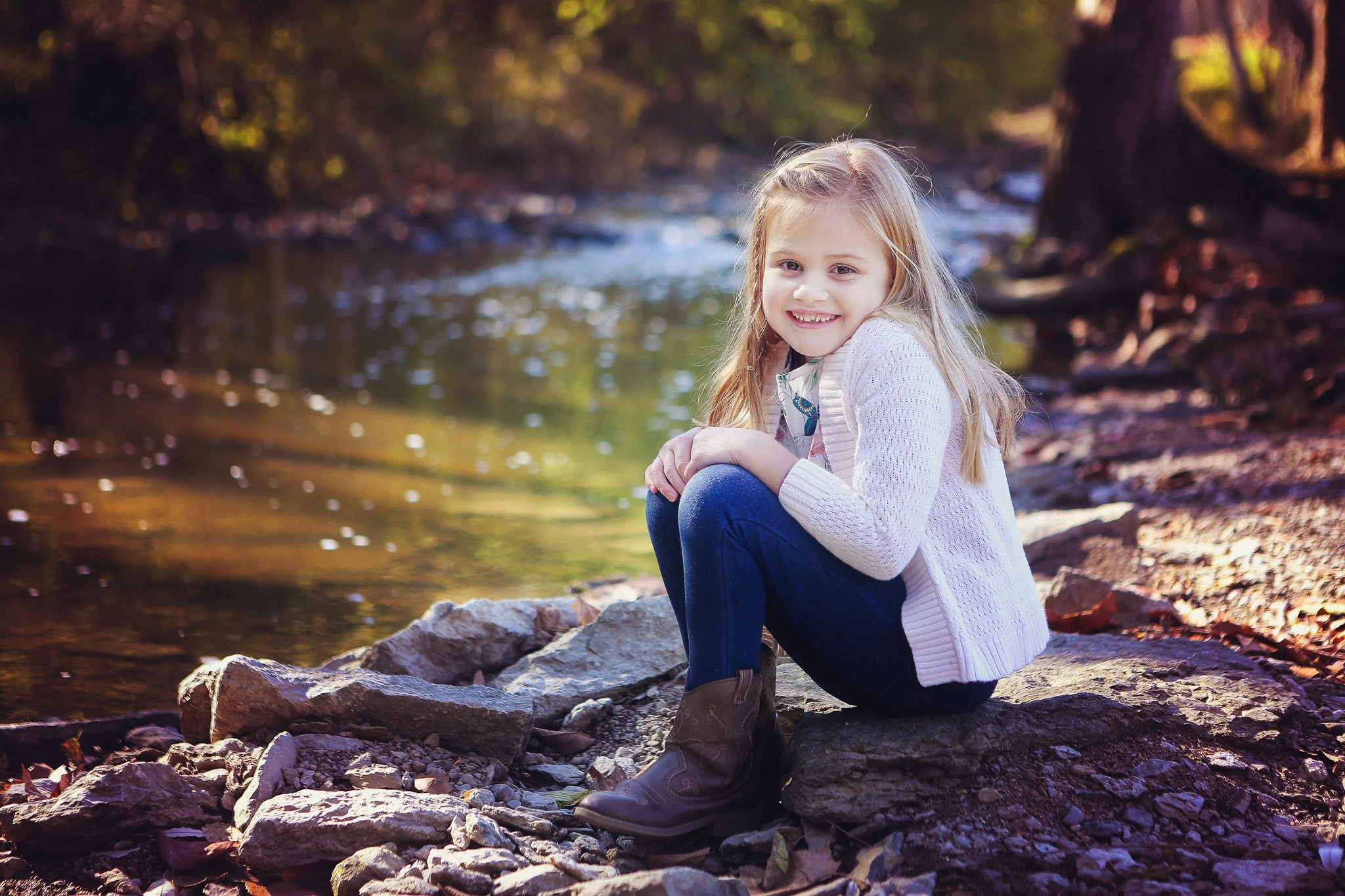 A young girl sitting on rocks by a river in a forest during autumn, smiling at the camera.