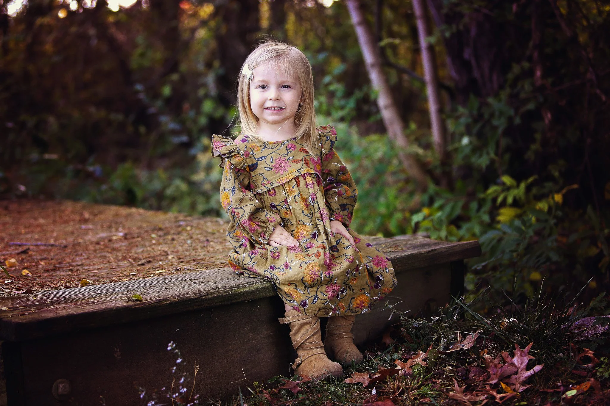 A young girl with blonde hair sitting on a wooden plank in a forested area, smiling at the camera.