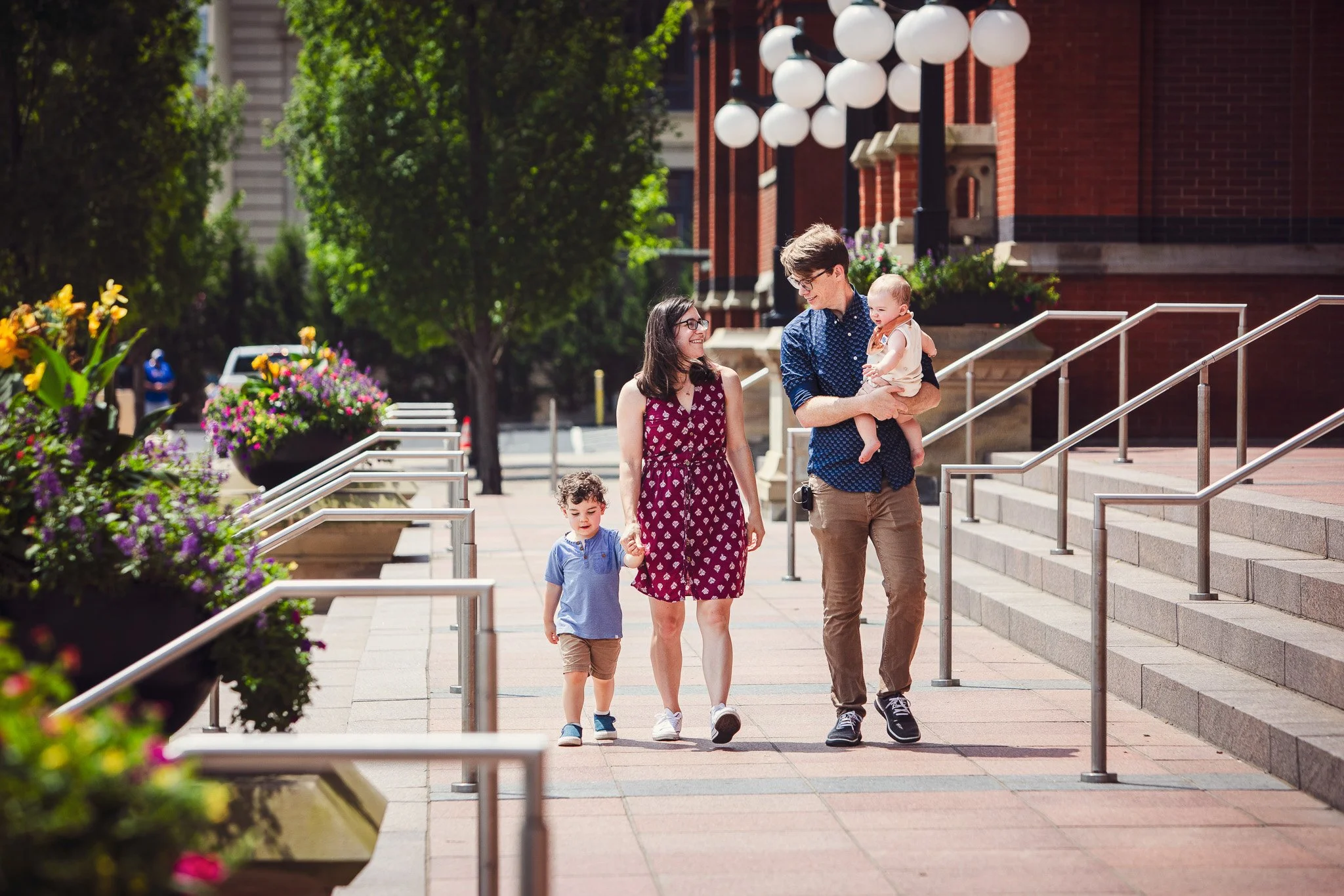 A family of four walking on a city sidewalk in front of a brick building with outdoor stairs and decorative street lamps, with flower planters nearby.