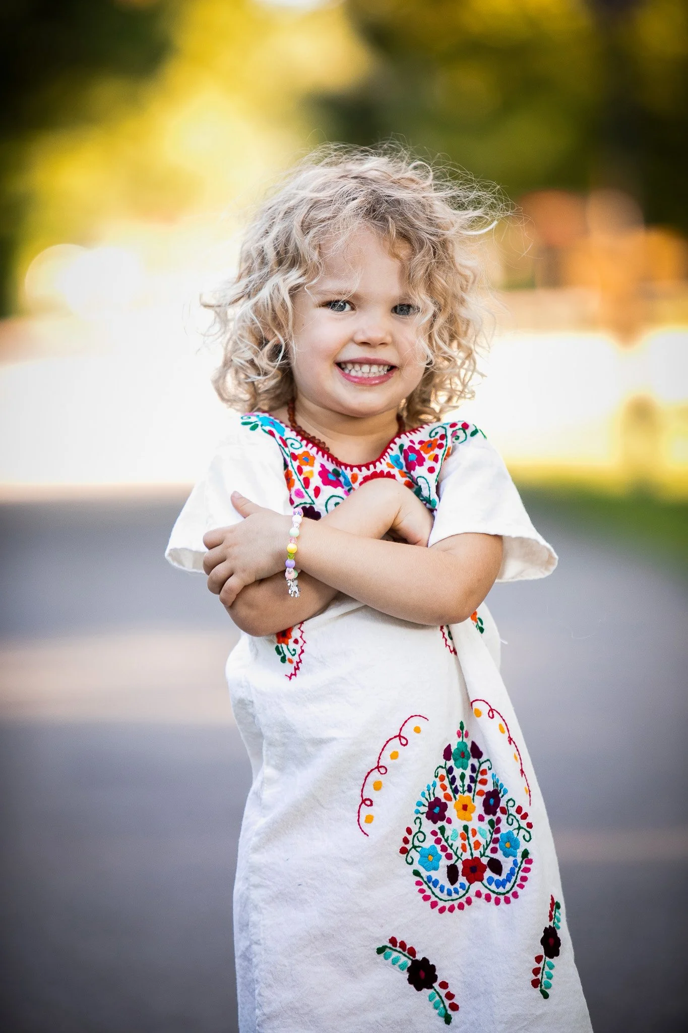 A young girl with curly blonde hair smiling, wearing a white embroidered dress with colorful floral patterns, standing outdoors on a sunny day.