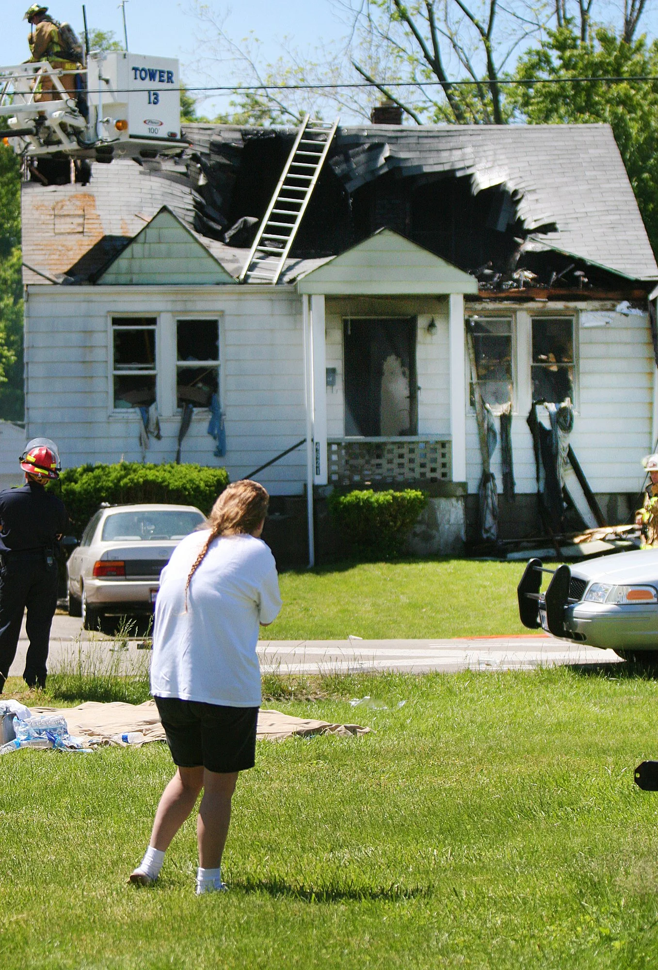 A house with extensive fire damage on the roof, firefighters and onlookers at the scene, with a person with long hair in a white shirt and black shorts in the foreground.