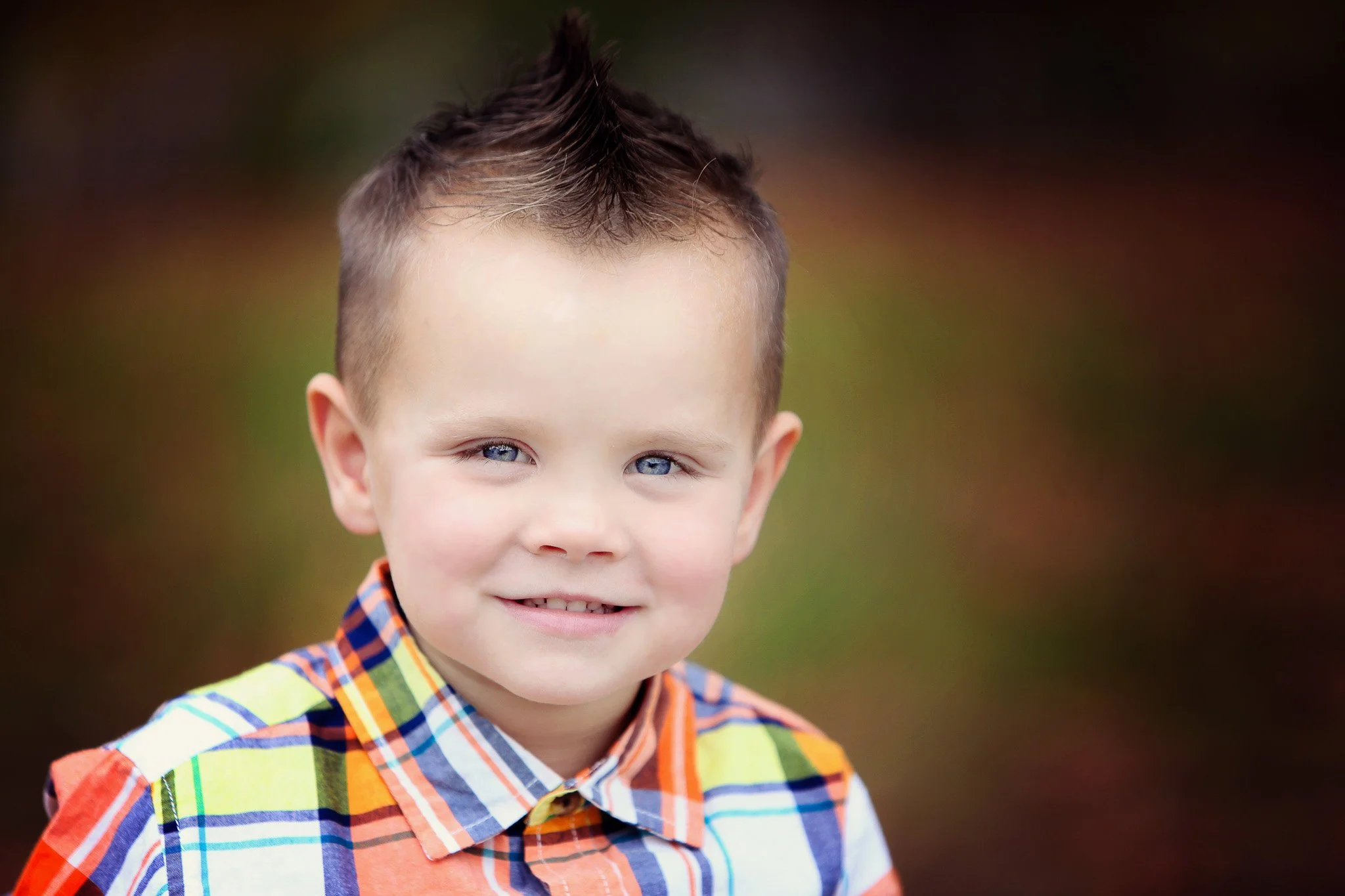Close-up of a smiling young boy with blue eyes, wearing a colorful plaid shirt, outdoors with blurred autumn background.