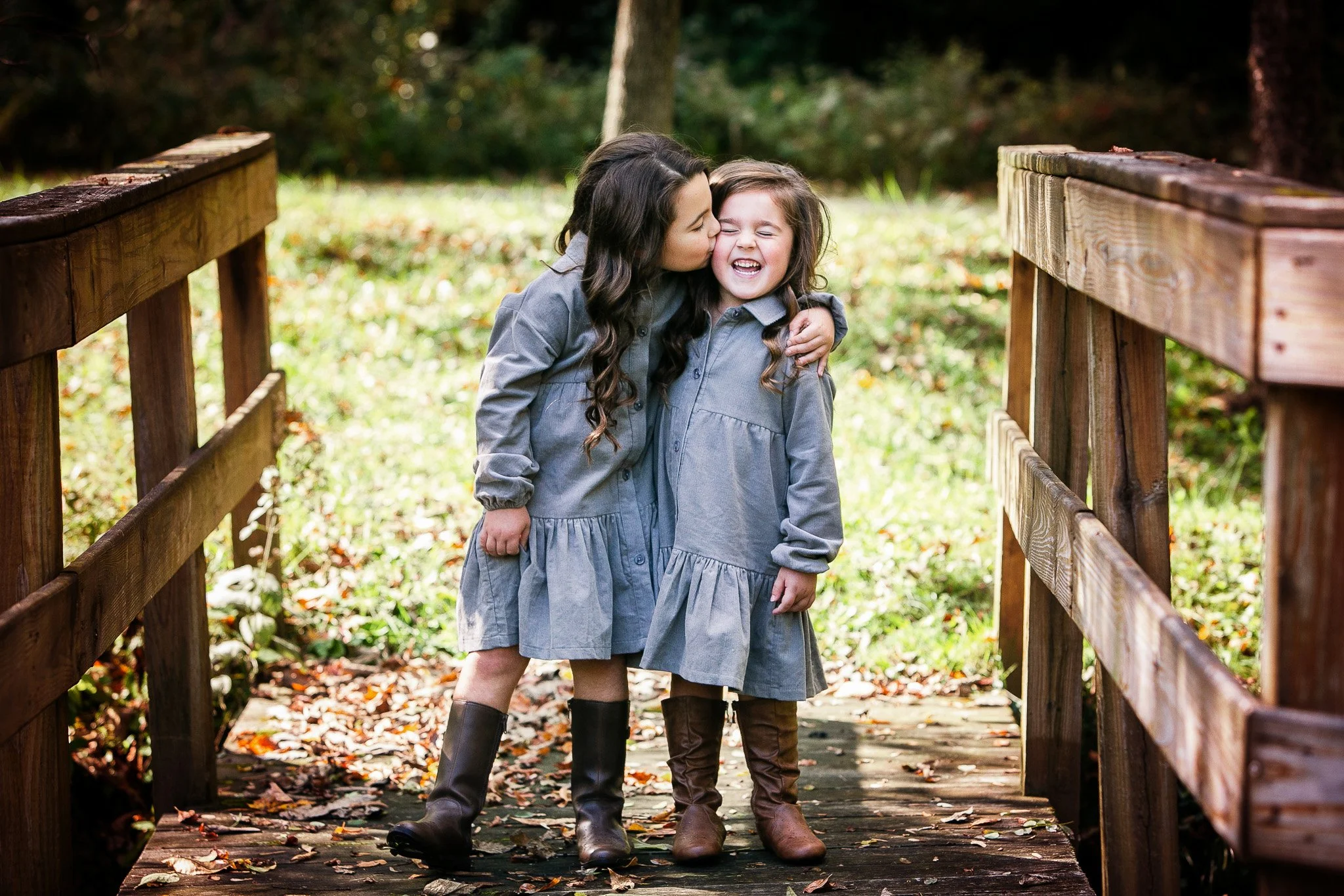 Two young girls in gray dresses and brown boots hugging and smiling on a wooden bridge in a park or forest area with fallen leaves.