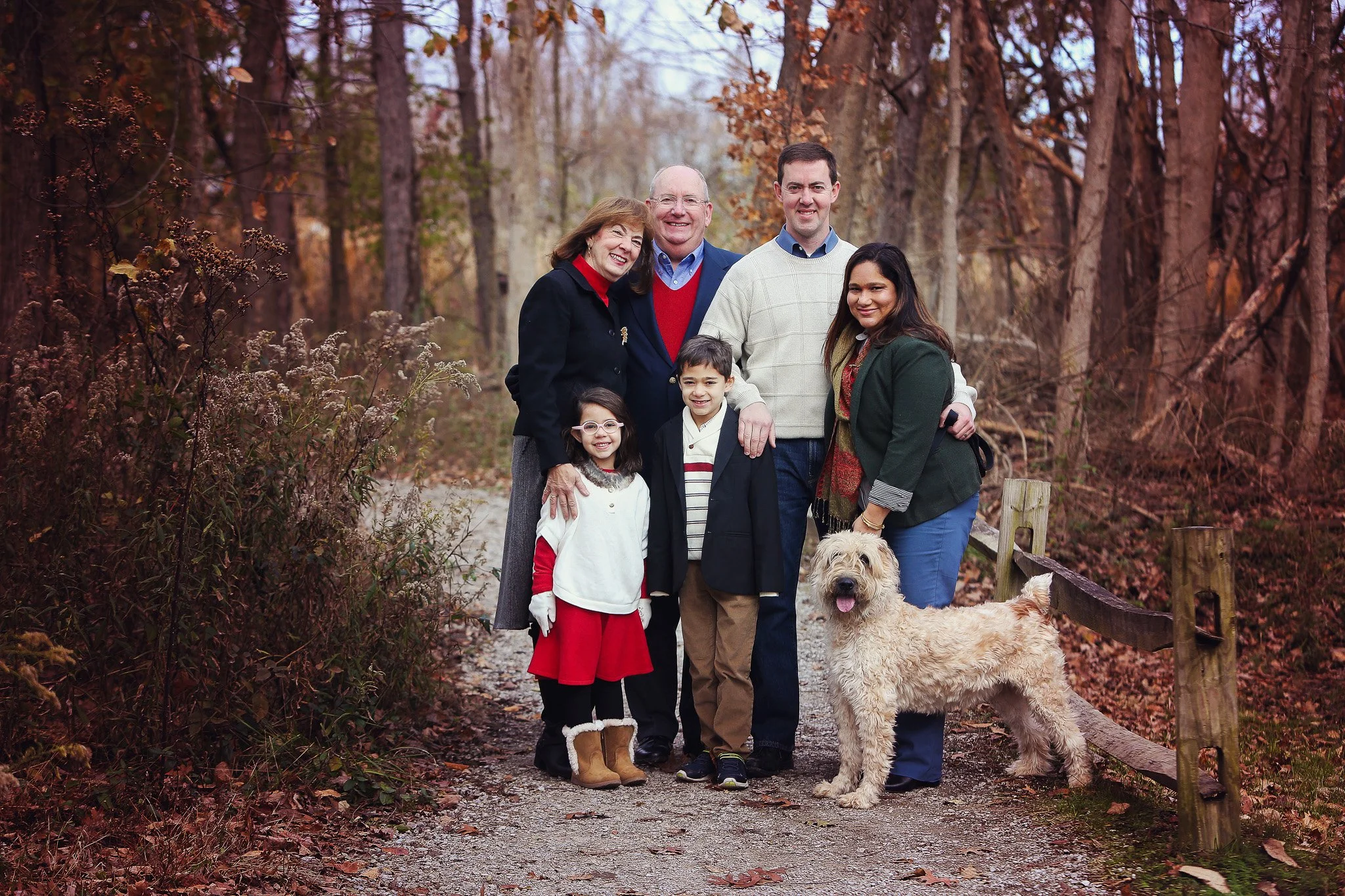 Family photo of seven people and a dog on a wooded trail during fall, with trees and fallen leaves in the background.