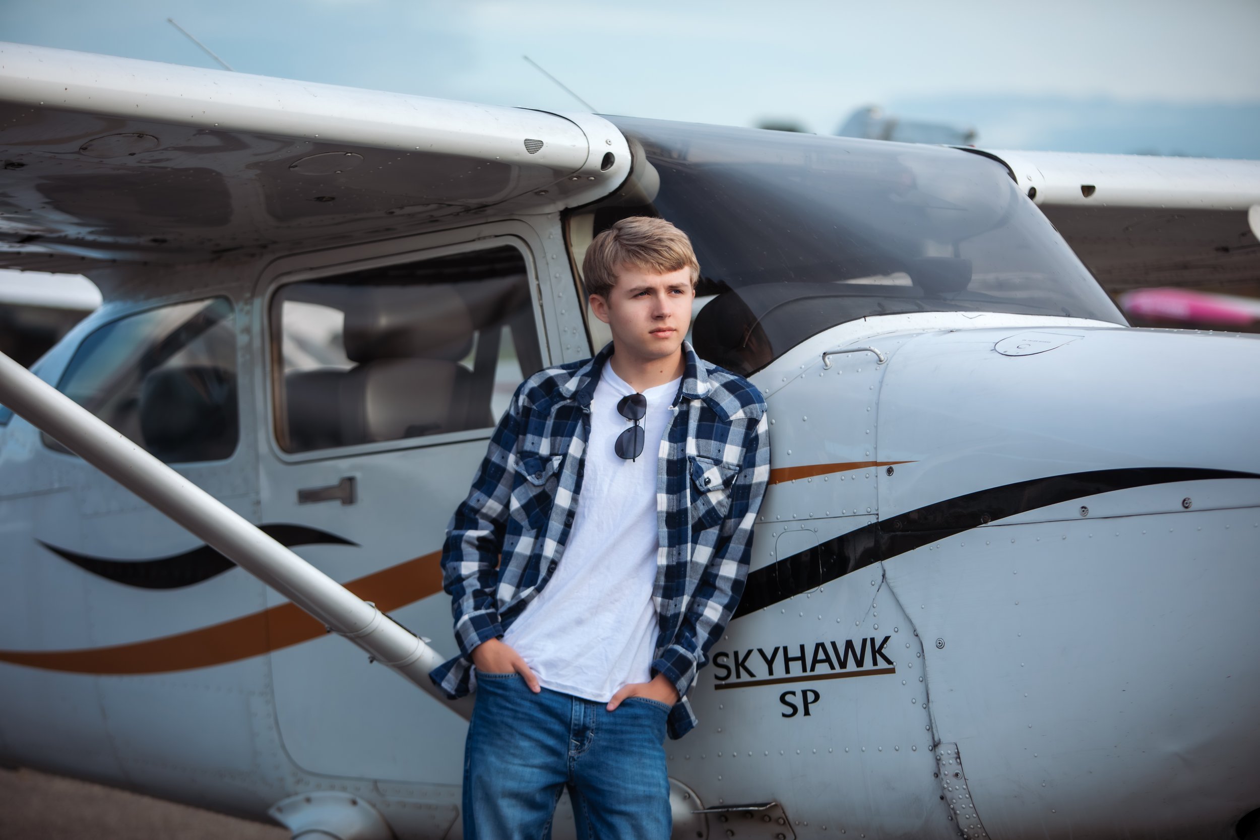 A teenage boy with blonde hair wearing a plaid shirt, white T-shirt, and jeans stands next to a small white airplane labeled 'Skyhawk SP,' with sunglasses hanging from his shirt collar, looking off to the side.