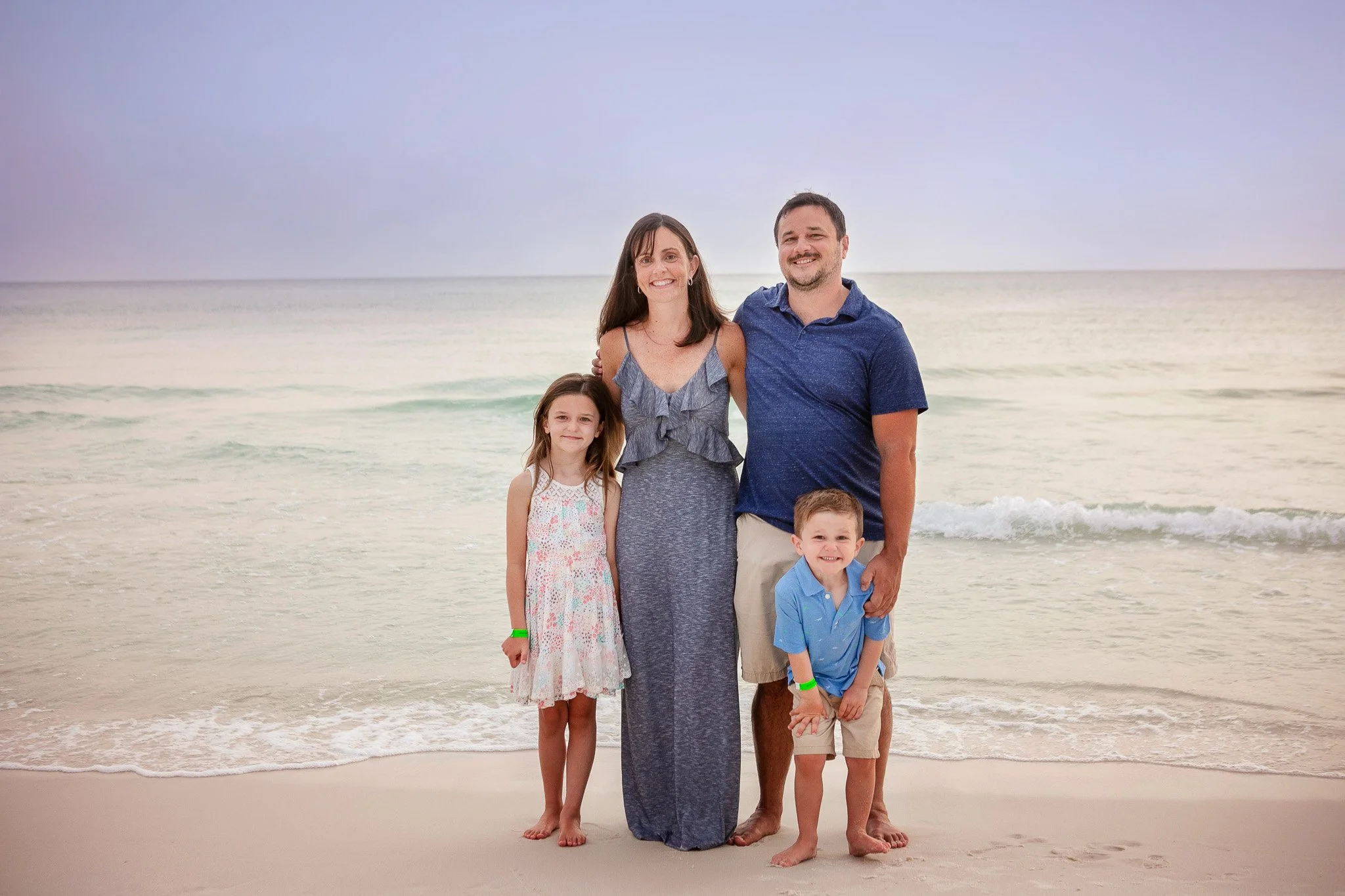 Family of four posing on the beach with the ocean in the background. The mother, father, daughter, and son are smiling and standing close together.