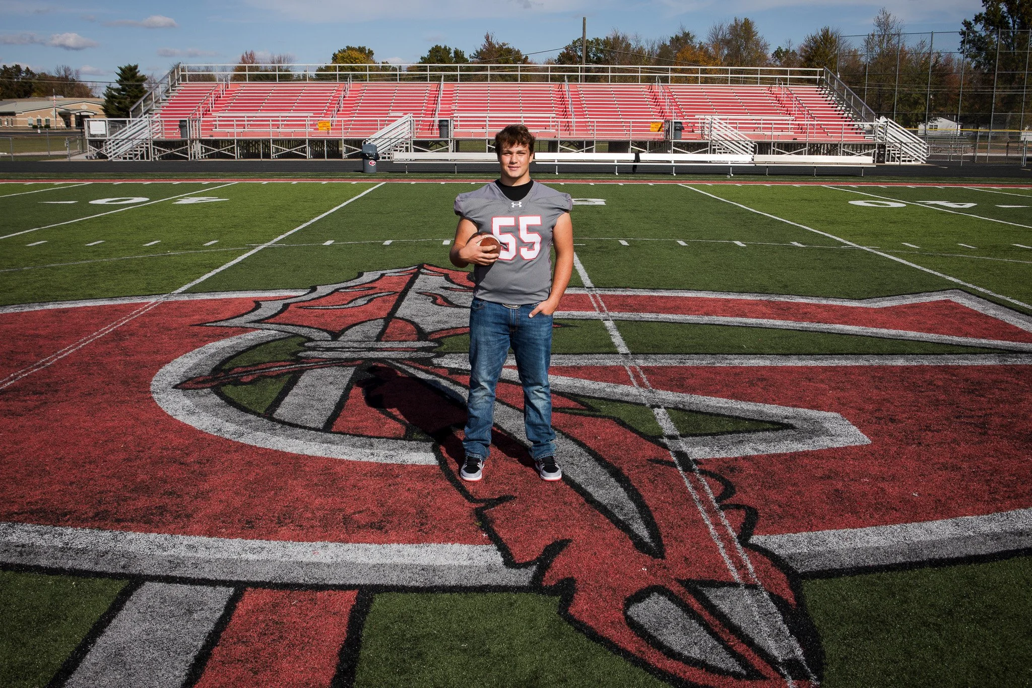 A young man holding a football standing on a football field with a large red cardinal logo in the center. He is wearing a football jersey with the number 55. The background includes bleachers and trees.